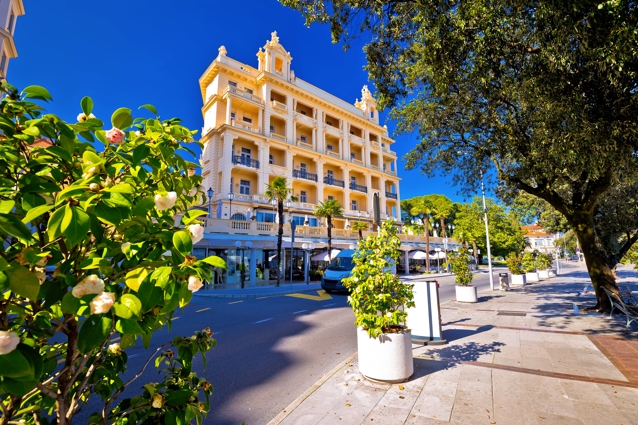Gelbes Hotel mit Balkonen an einer von Bäumen gesäumten Straße bei blauem Himmel.