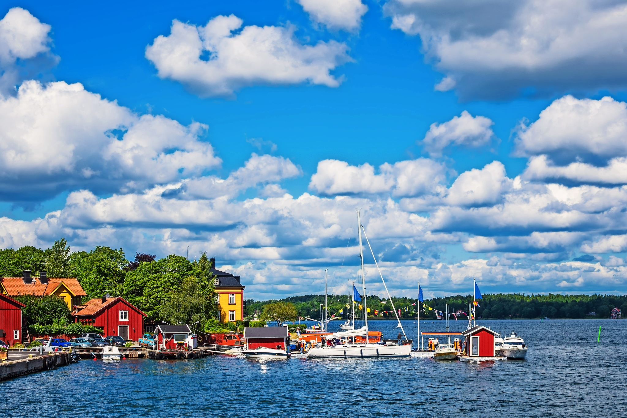 Ein malerischer Hafen an der Küste mit roten Holzhäusern, mehreren Segelbooten im Wasser und einer grünen und waldreichen Landschaft im Hintergrund unter einem blauen Himmel mit weißen Wolken.