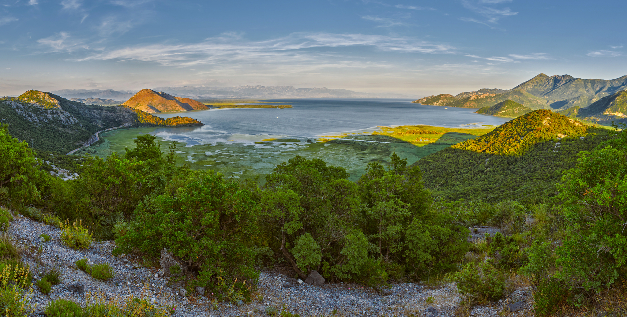 Panorama eines Sees mit umliegenden grünen Hügeln und blauem Himmel.