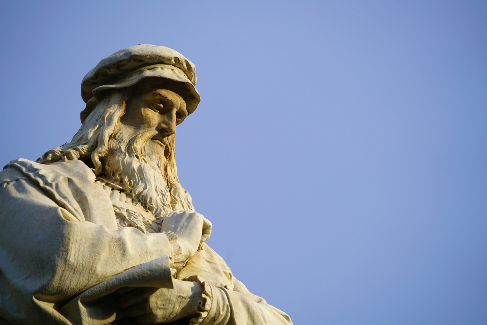 Steinfigur eines nachdenklichen Mannes mit Bart und Mütze vor blauem Himmel.