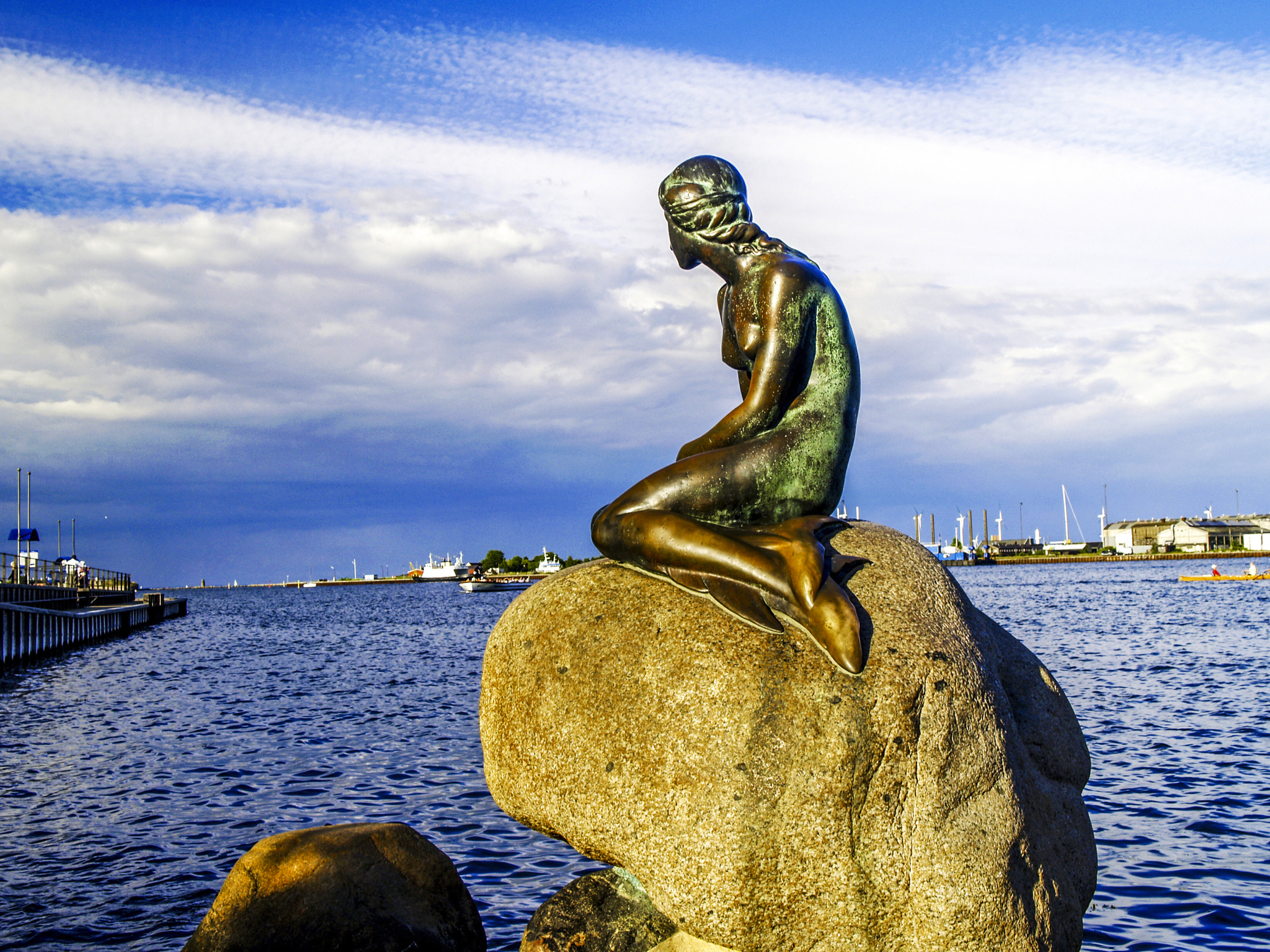 Eine Bronzestatue einer Meerjungfrau sitzt auf einem großen Felsen am Ufer eines Gewässers unter einem blauen Himmel mit einigen Wolken. Im Hintergrund sind Boote und Gebäude zu sehen.