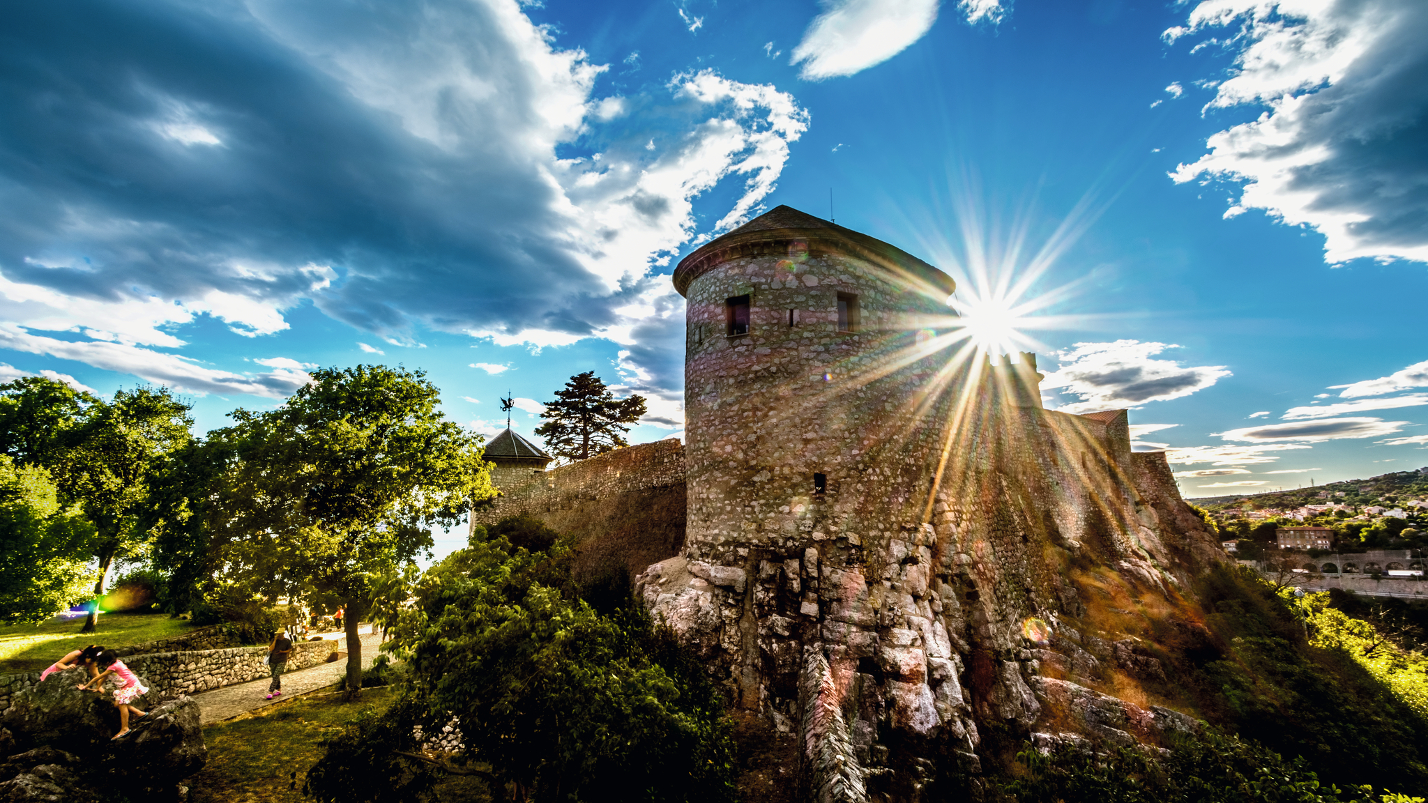 Alte Steinburg mit Sonnenstrahlen, umgeben von Bäumen und blauen Himmel.