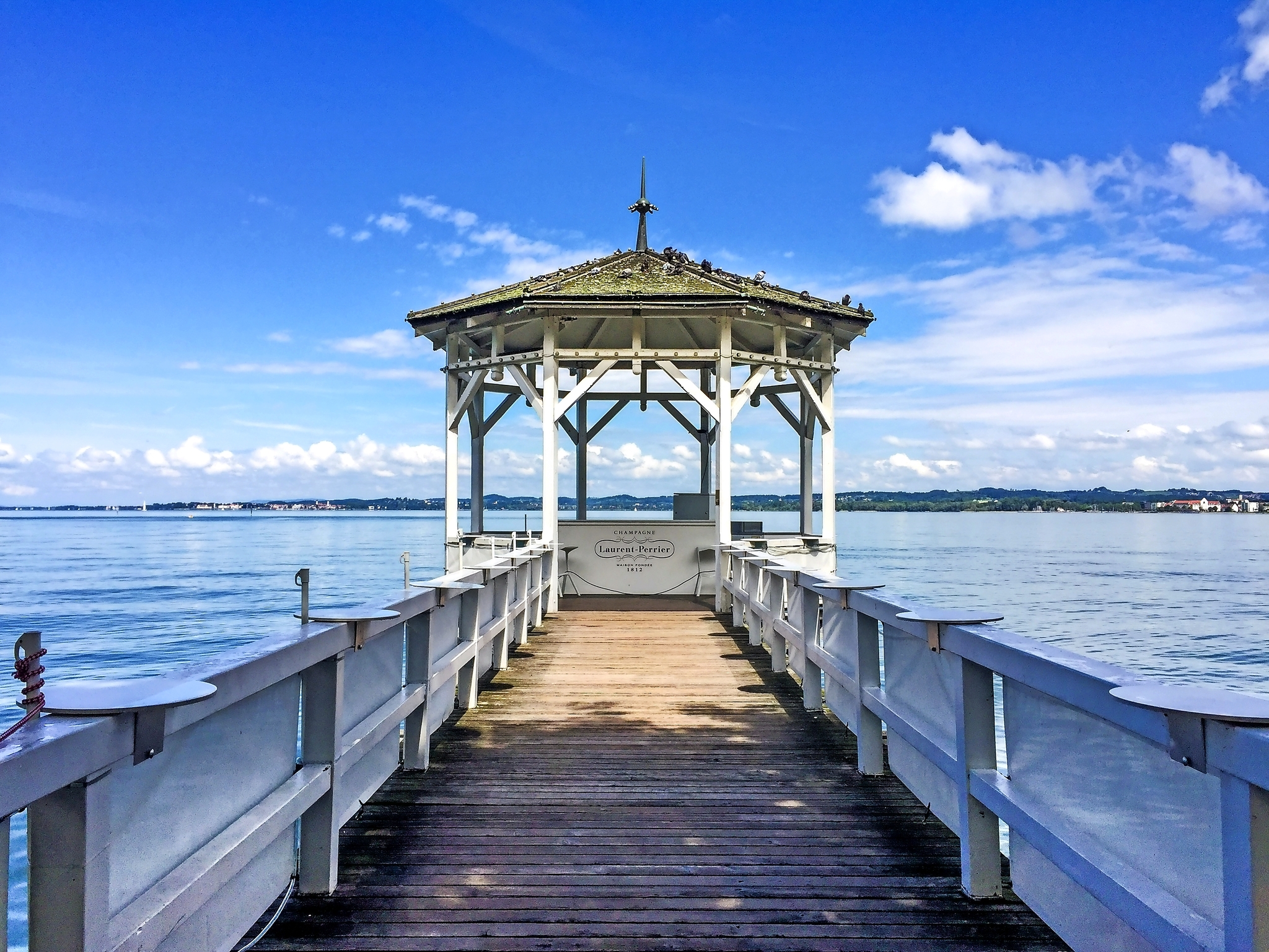 Holzsteg, der zu einem Pavillon über einem See führt, unter blauem Himmel mit weißen Wolken.