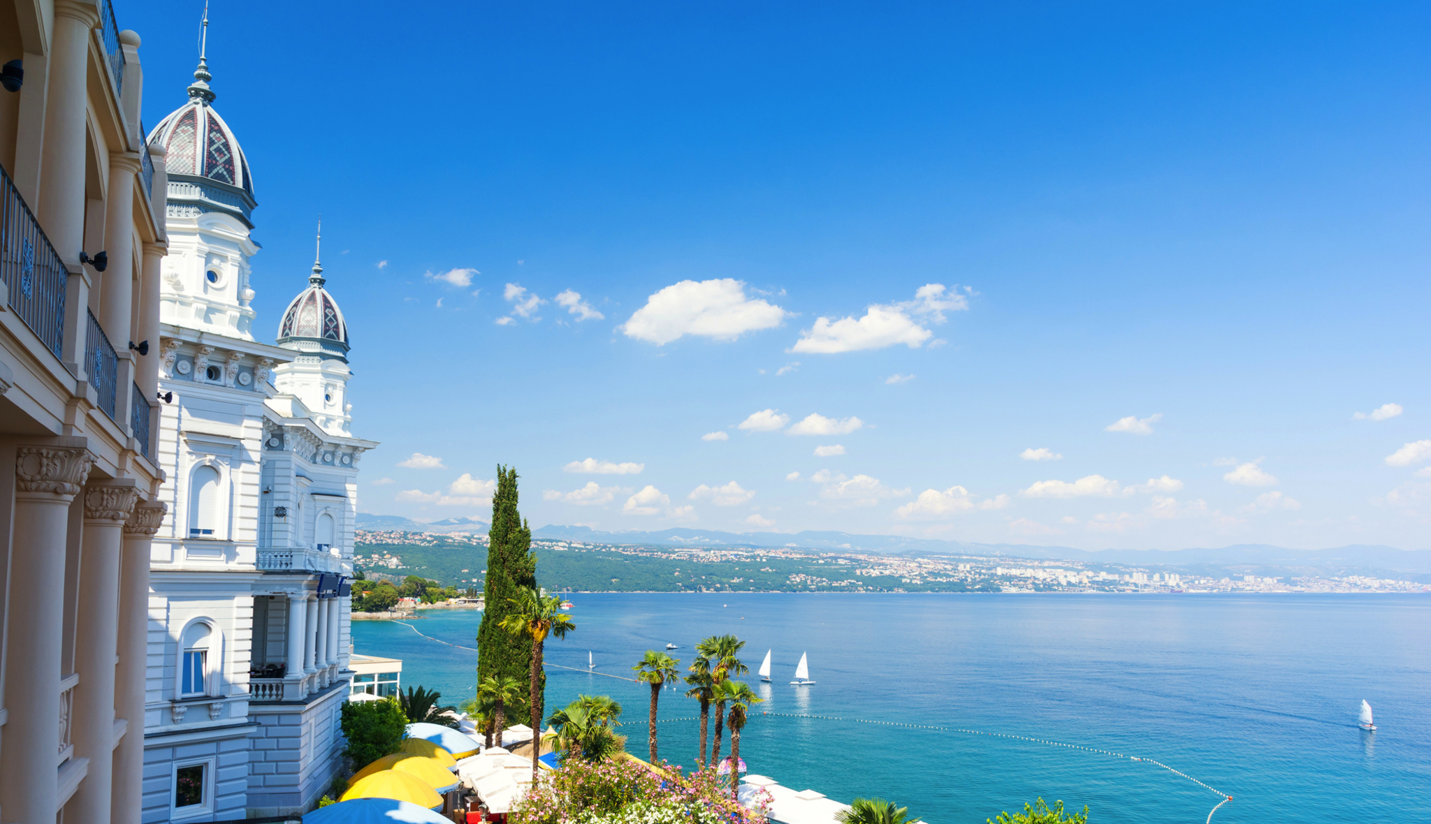 Küstenlandschaft mit historischem Gebäude, Palmen und Segelbooten auf blauem Meer.