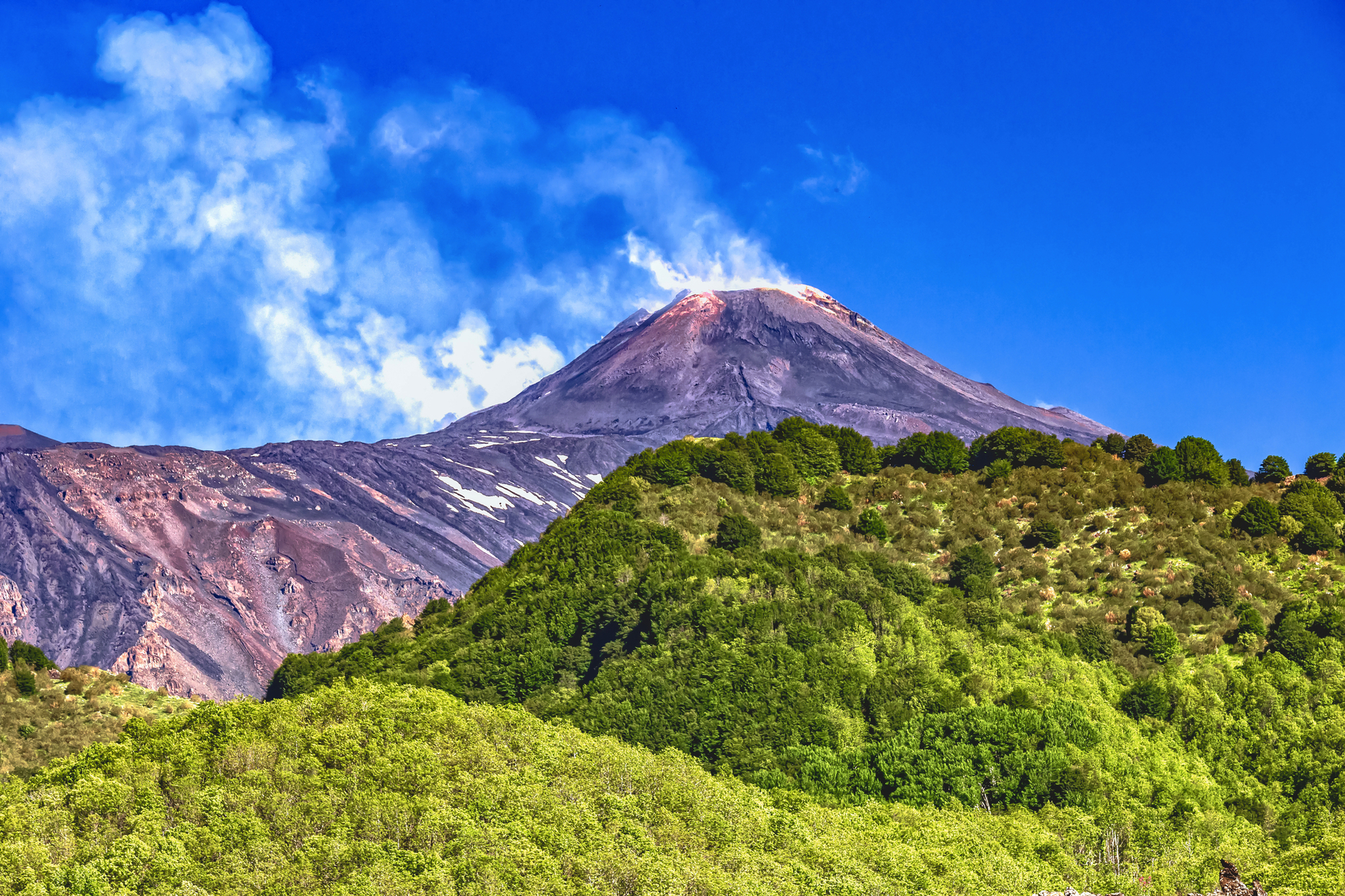 Vulkan mit rauchendem Gipfel, umgeben von grün bewaldeten Hügeln unter klarem blauen Himmel.