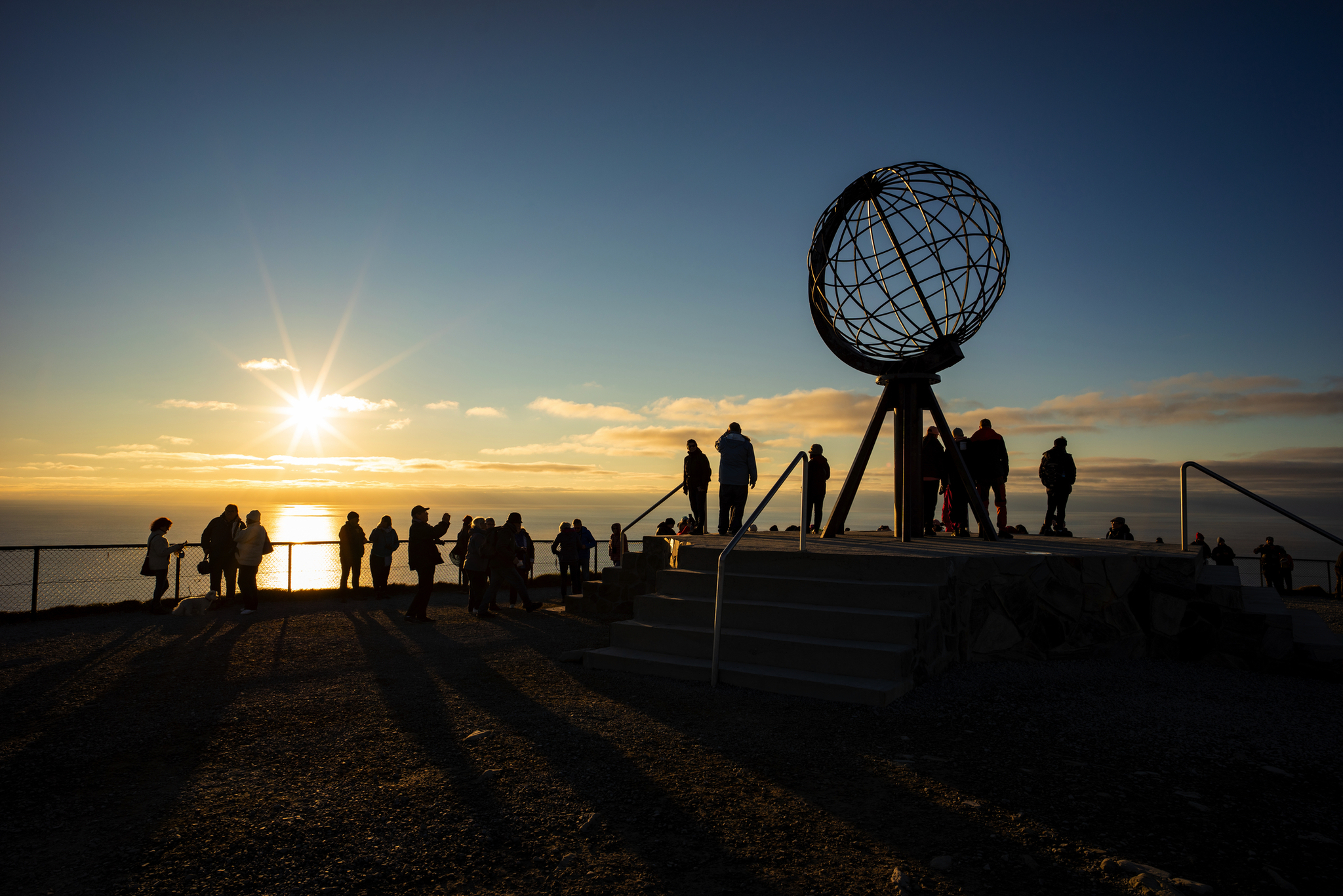 Sonnenuntergang am Nordkap mit Globus-Skulptur und silhouettenhaften Menschen.