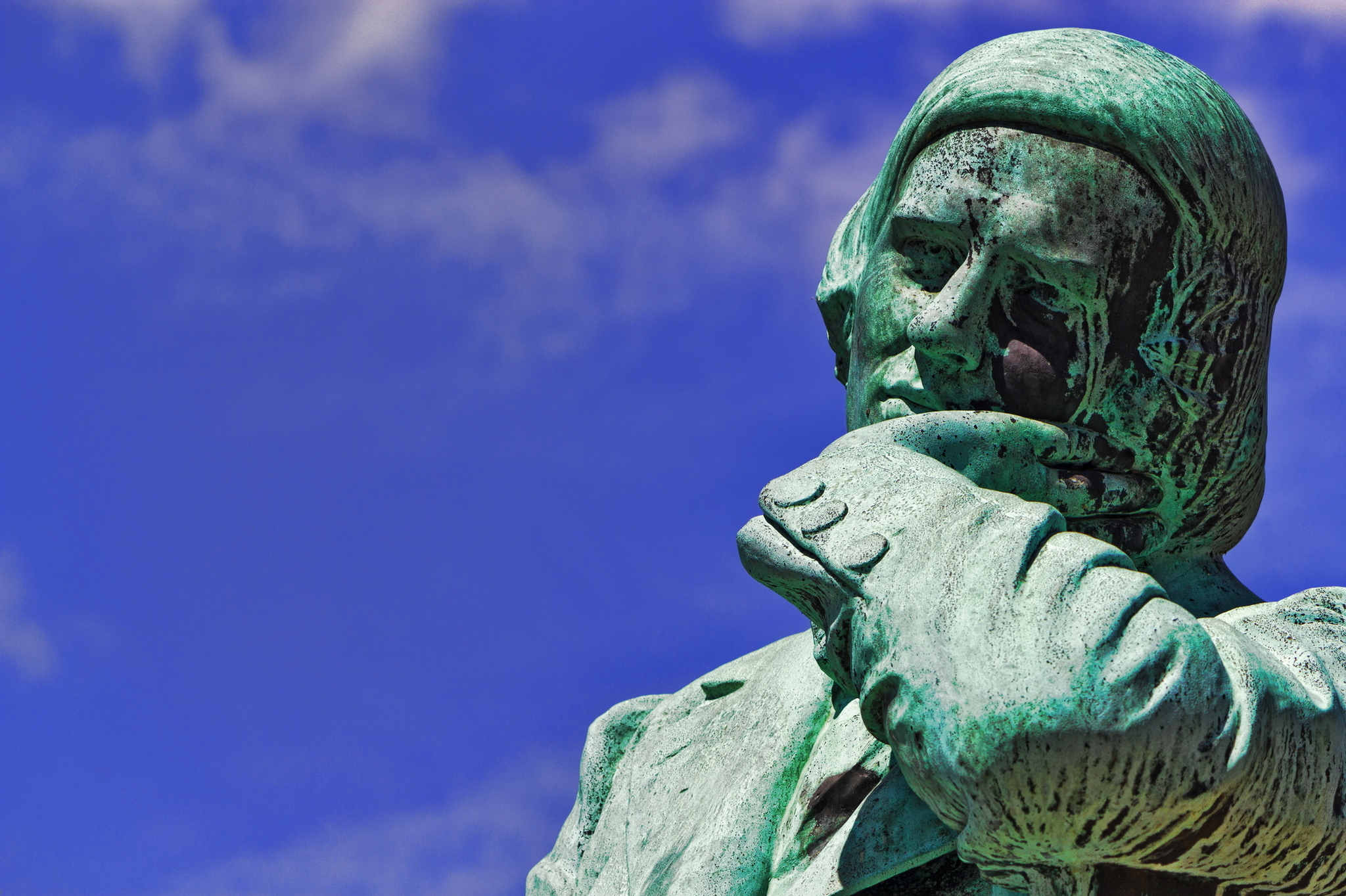 Grüne Statue eines nachdenklichen Mannes aus Bronze vor blauem Himmel.