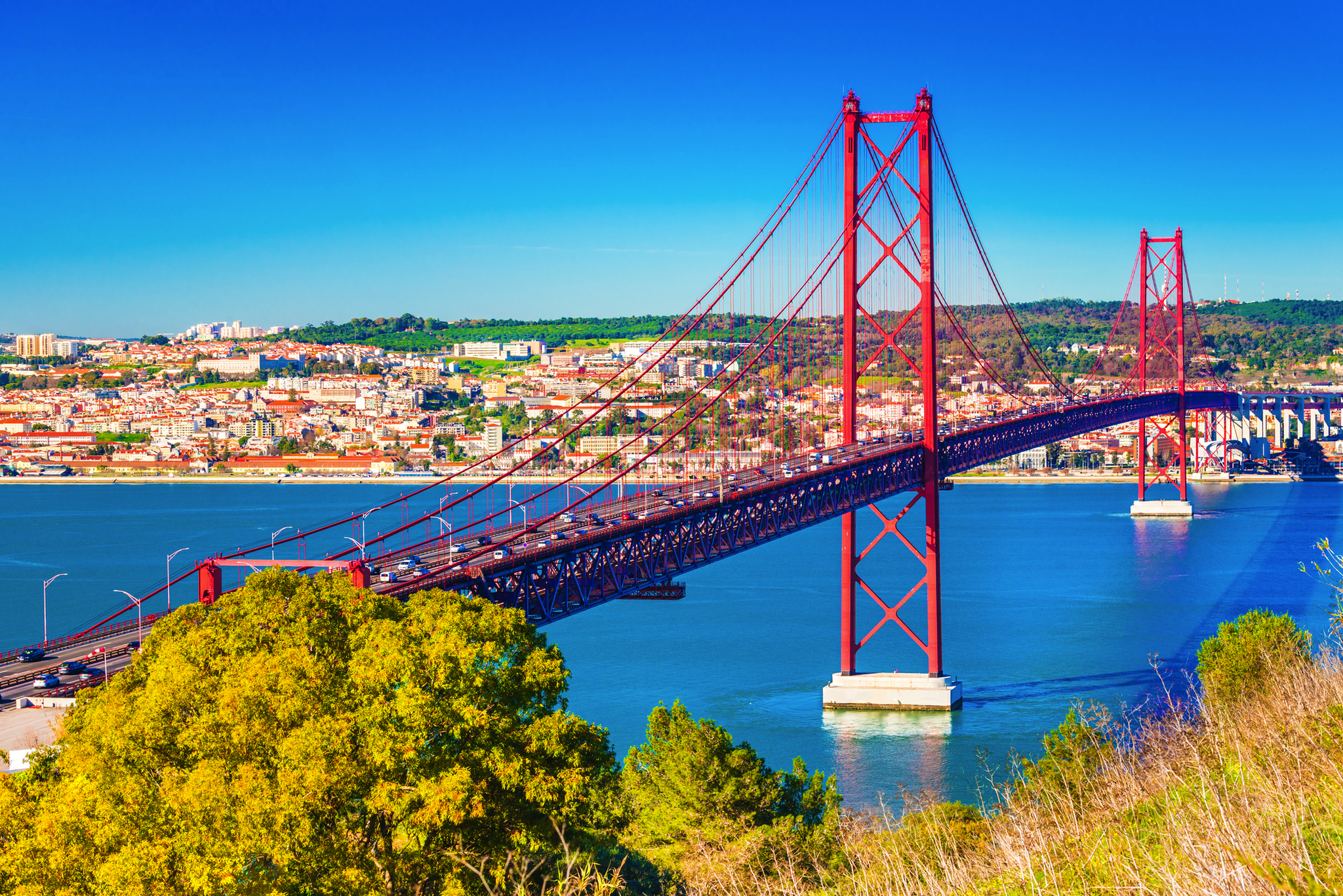 Rote Hängebrücke über einem Fluss mit Stadtansicht im Hintergrund.