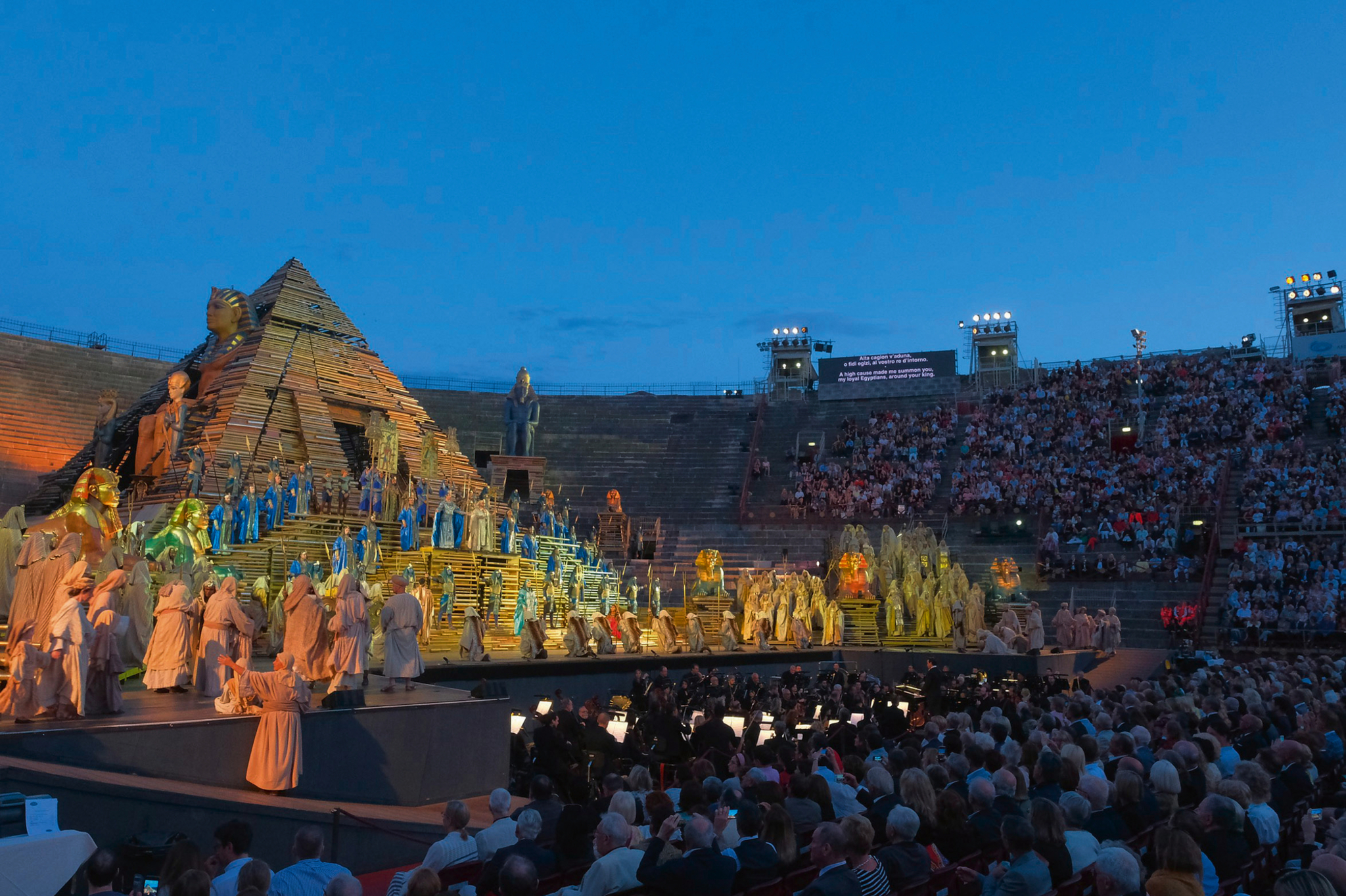 Bühnenaufführung von Verdis Aida in antikem Stadion mit Pyramide und Publikum.