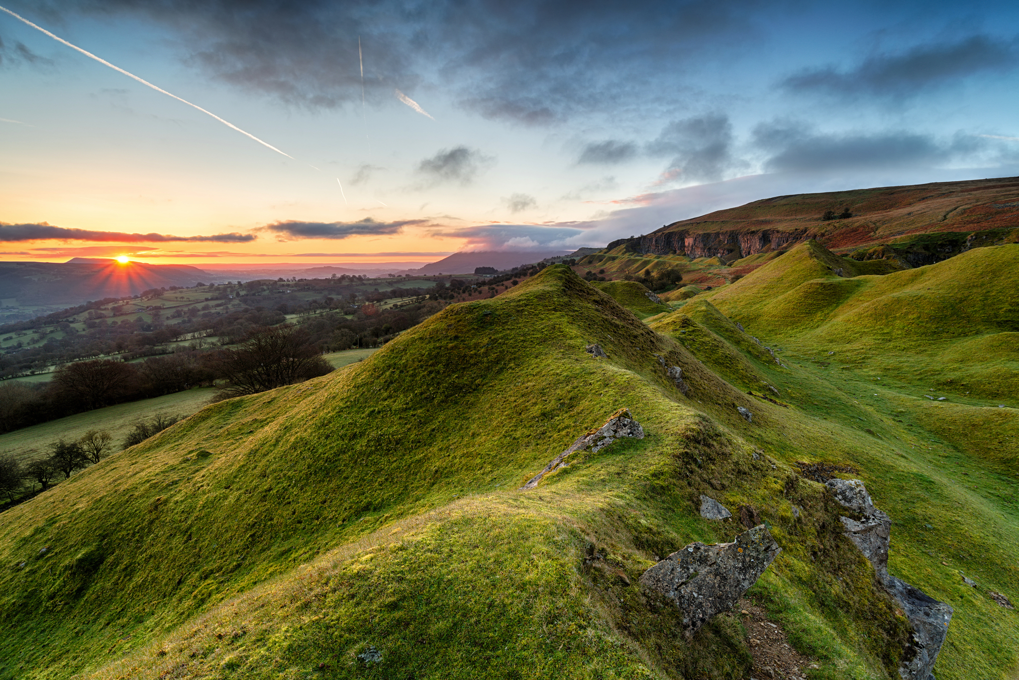 Hügelige Landschaft bei Sonnenuntergang mit bewölktem Himmel und Felsen.
