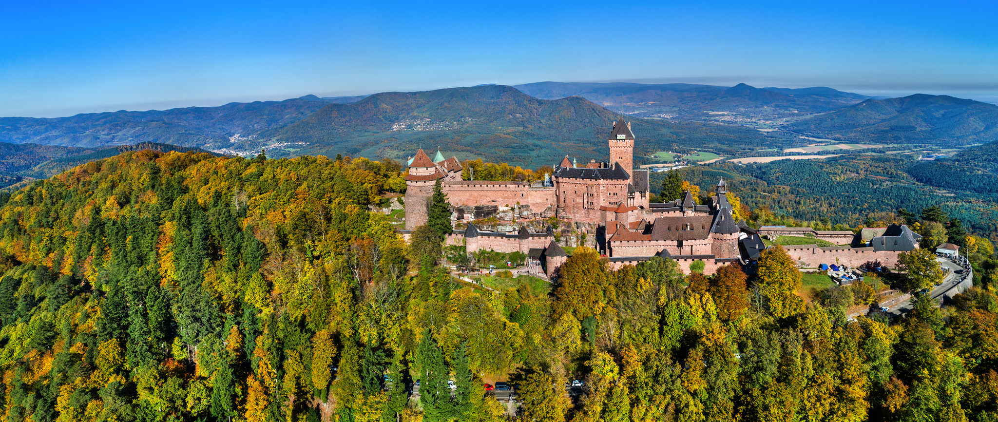 Luftaufnahme einer großen, alten Burg auf einem bewaldeten Hügel in herbstlicher Landschaft.