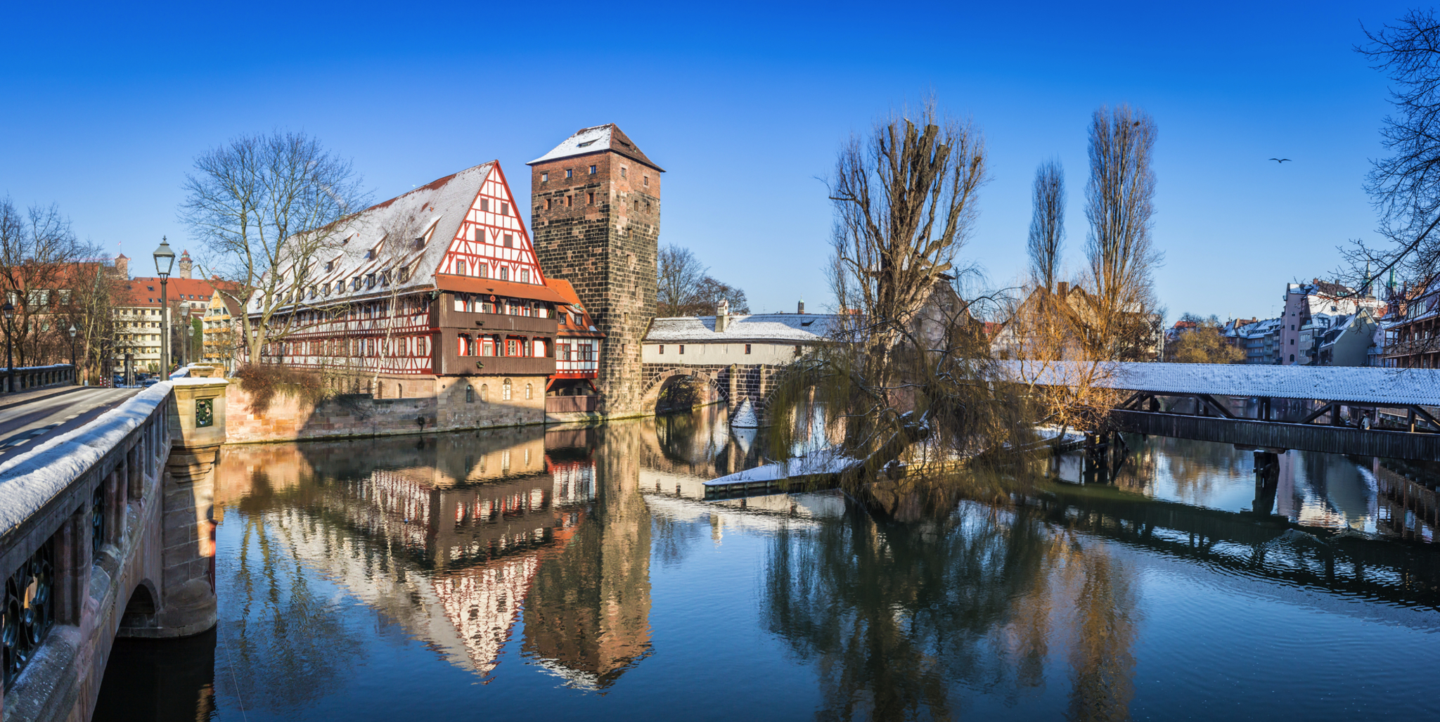 Fachwerkhaus und Brücke an verschneitem Flussufer, klarer Himmel, spiegelndes Wasser.