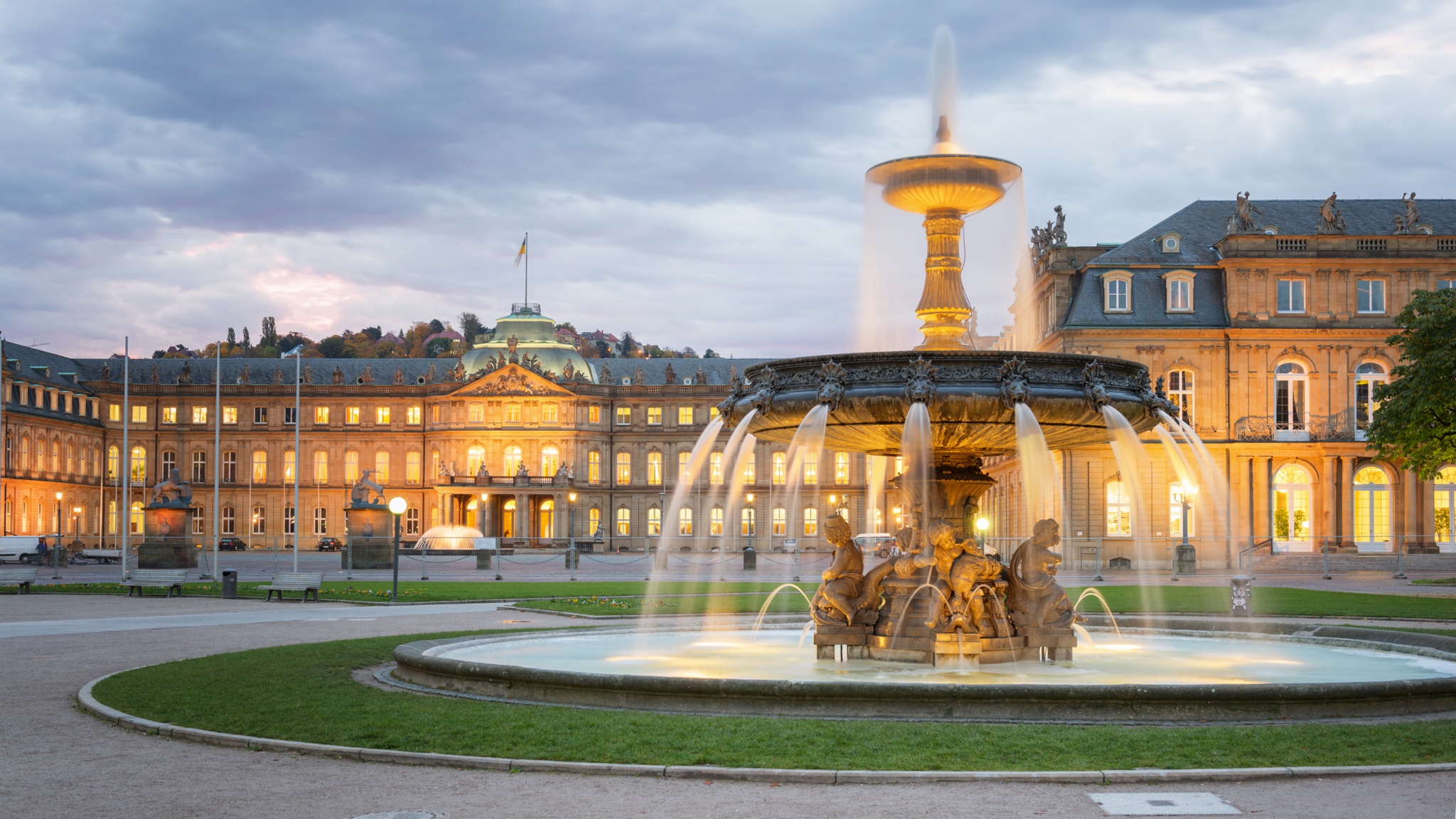Brunnen vor einem beleuchteten Schloss bei bewölktem Himmel.