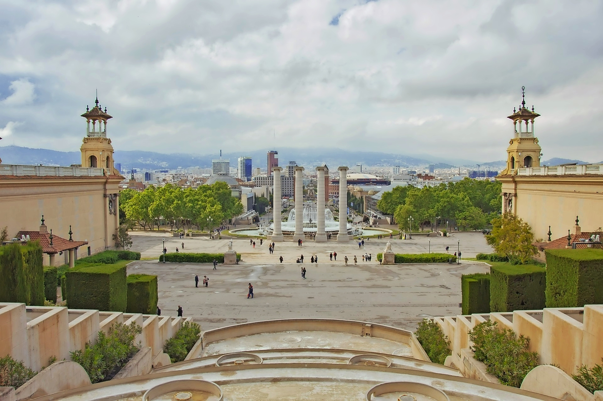 Blick von einer erhöhten Plattform auf eine große, mit Säulen verzierte Freitreppe, die zu einem offenen Platz führt, umgeben von Bäumen und historischen Gebäuden, mit einer Stadt im Hintergrund, unter einem bewölkten Himmel.