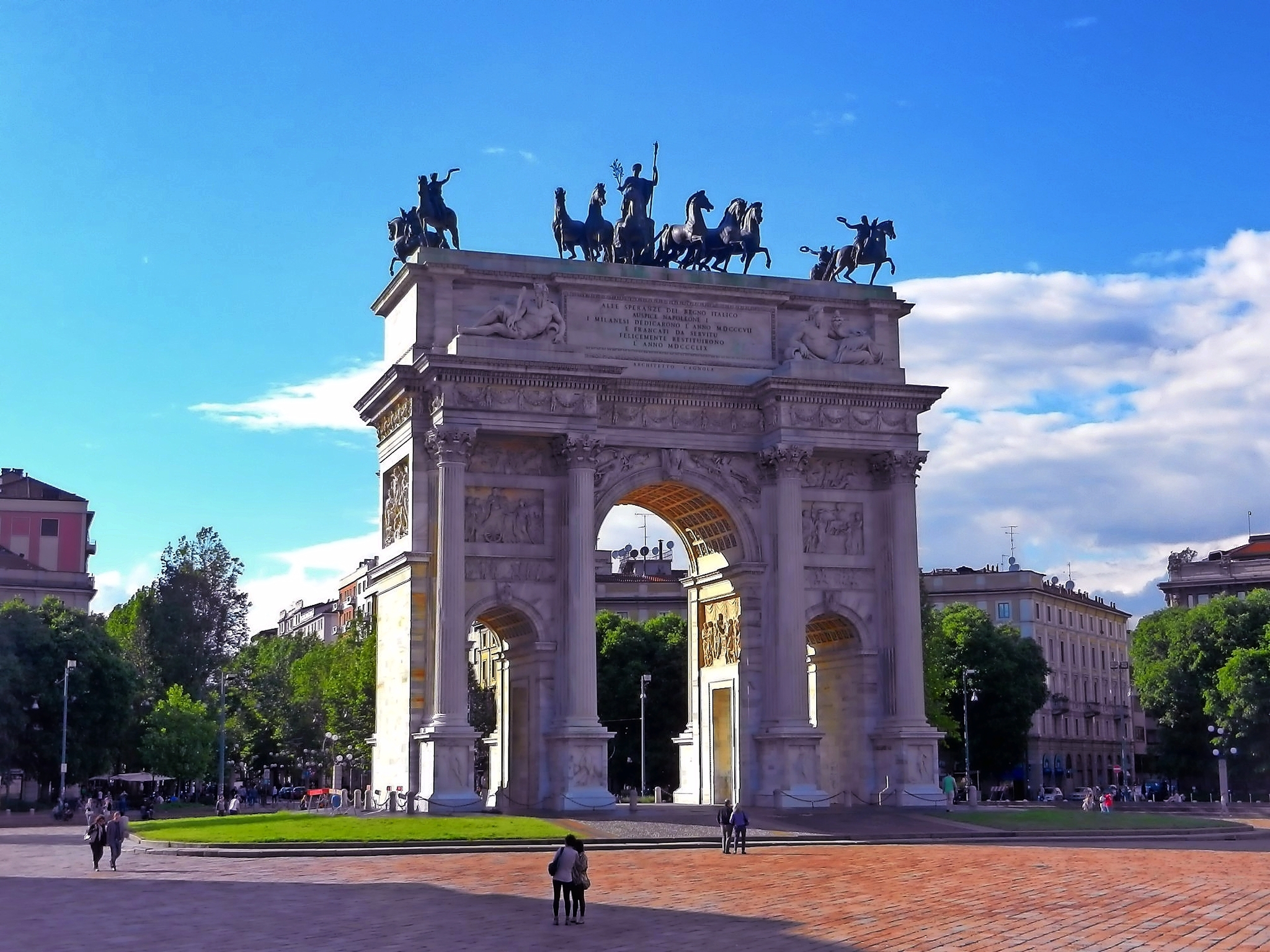 Triumphbogen mit Skulpturen auf Platz vor blauem Himmel.