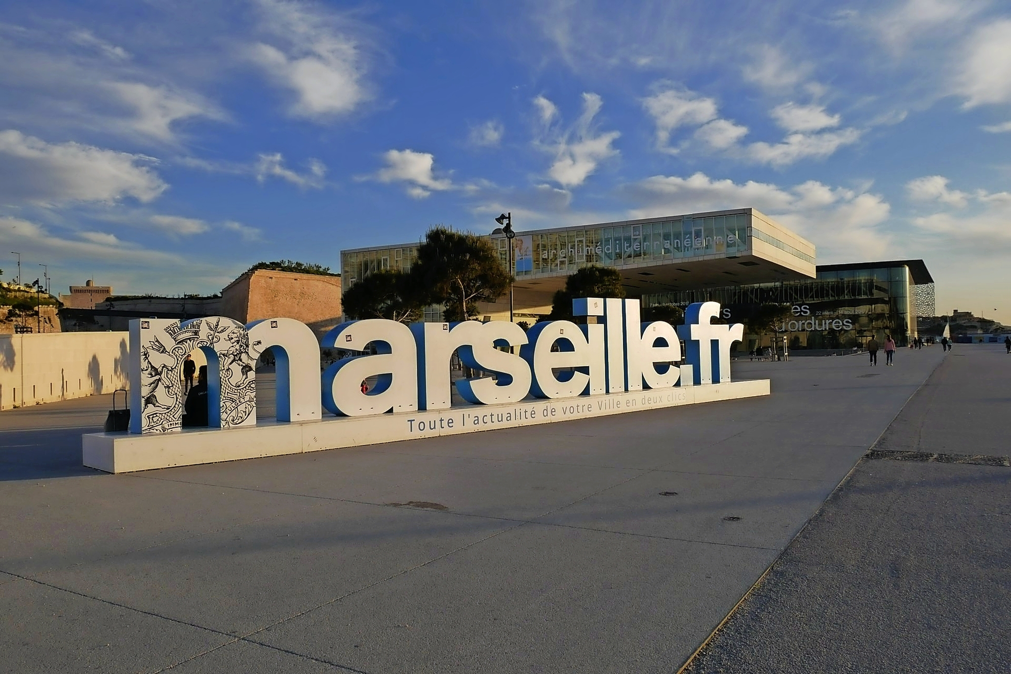 Große Schriftzüge "marseille.fr" vor modernem Gebäudekomplex bei Sonnenschein.