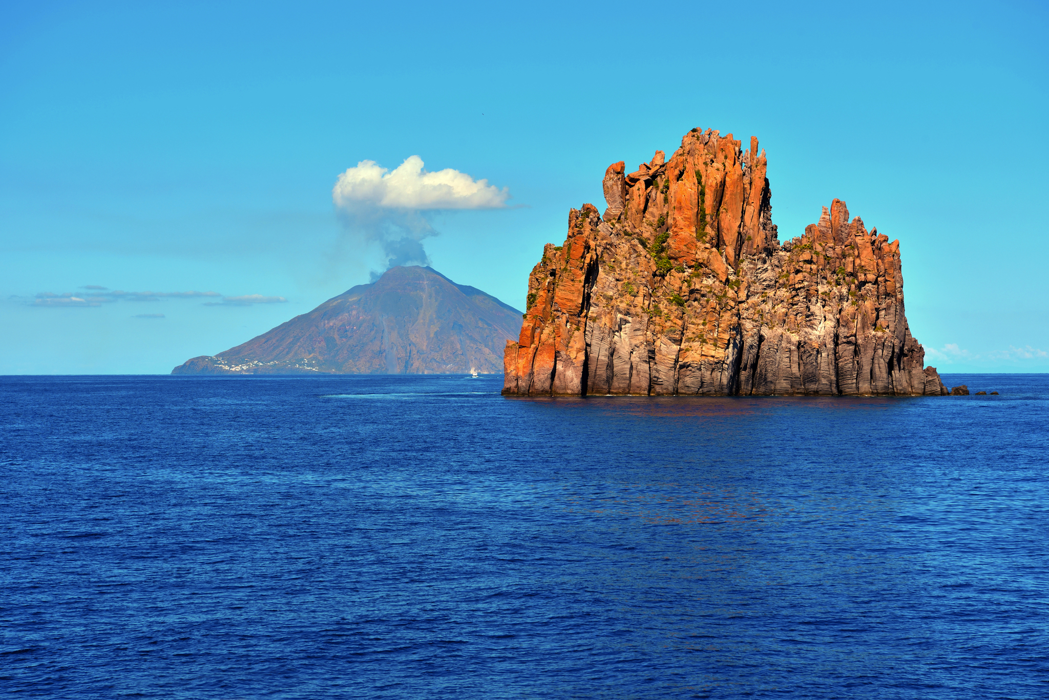Felsformation im Meer mit einem rauchenden Vulkan im Hintergrund unter blauem Himmel.