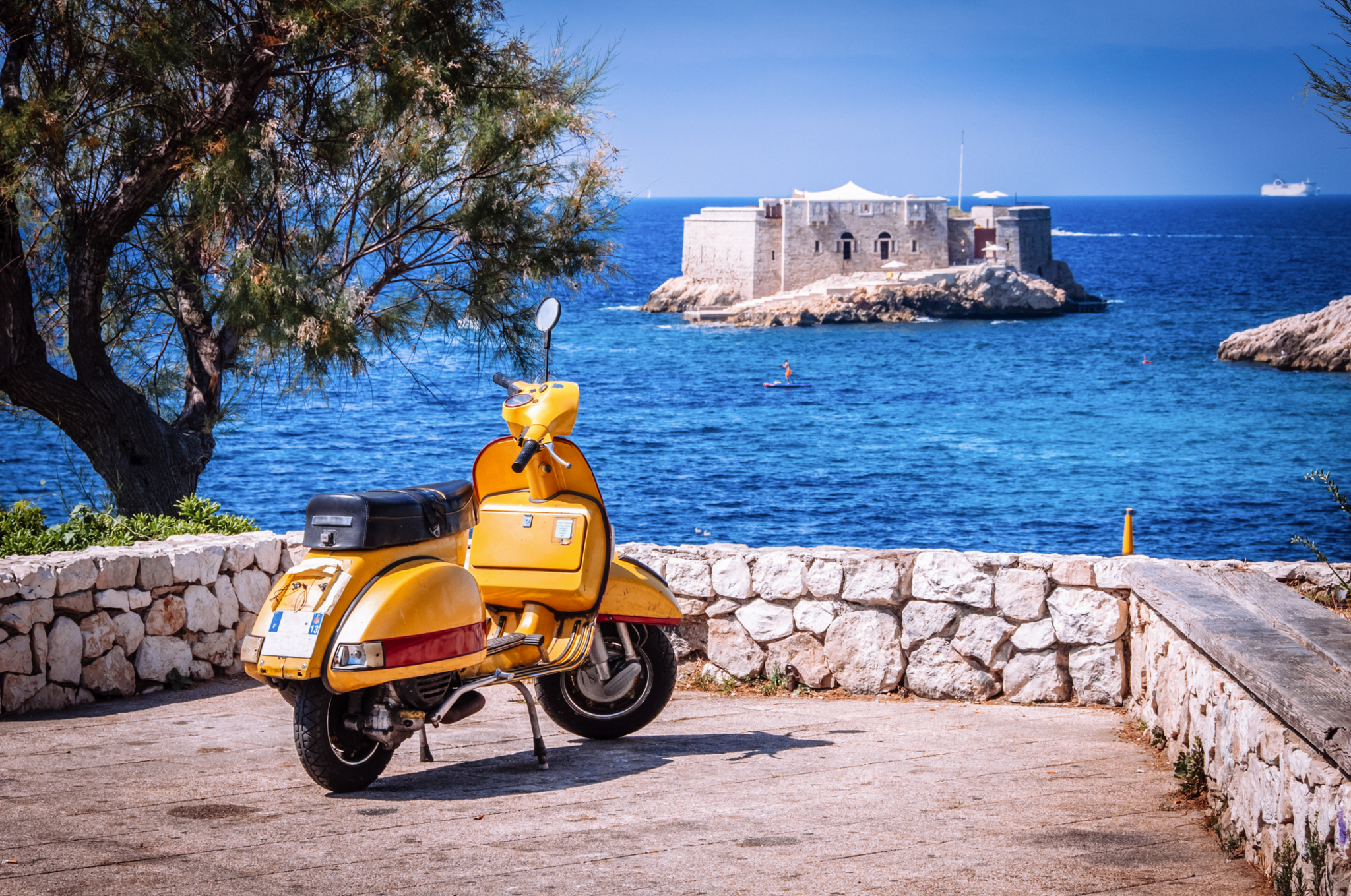 Gelber Motorroller vor Meer und Insel mit Festung im Hintergrund.