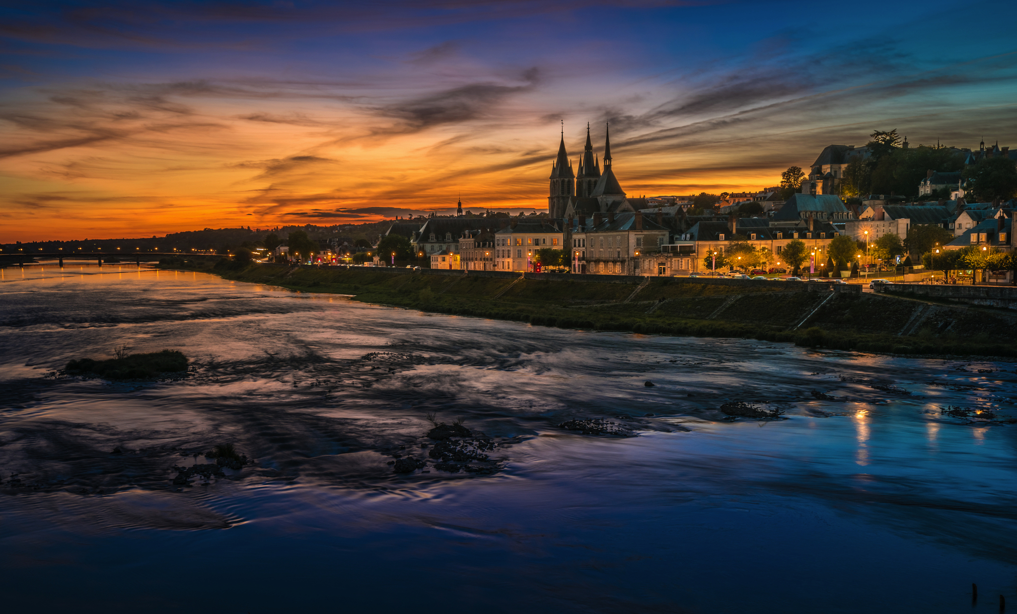 Flusslandschaft bei Sonnenuntergang mit beleuchteter Stadt und Kirche im Hintergrund.