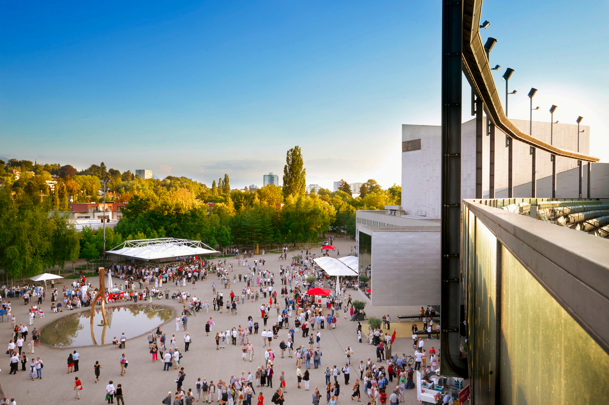 Menschenmenge auf einem Platz neben einem modernen Gebäude im Sonnenschein.