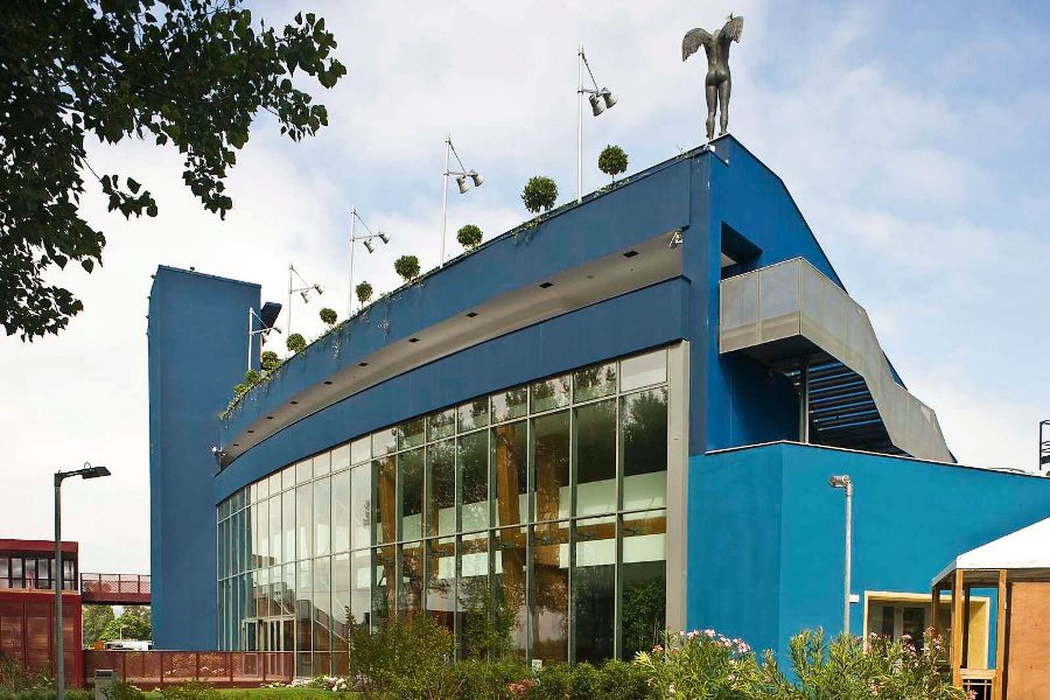 Blaues Gebäude mit Glasfassade, Bäumen auf dem Dach und einer Statue.