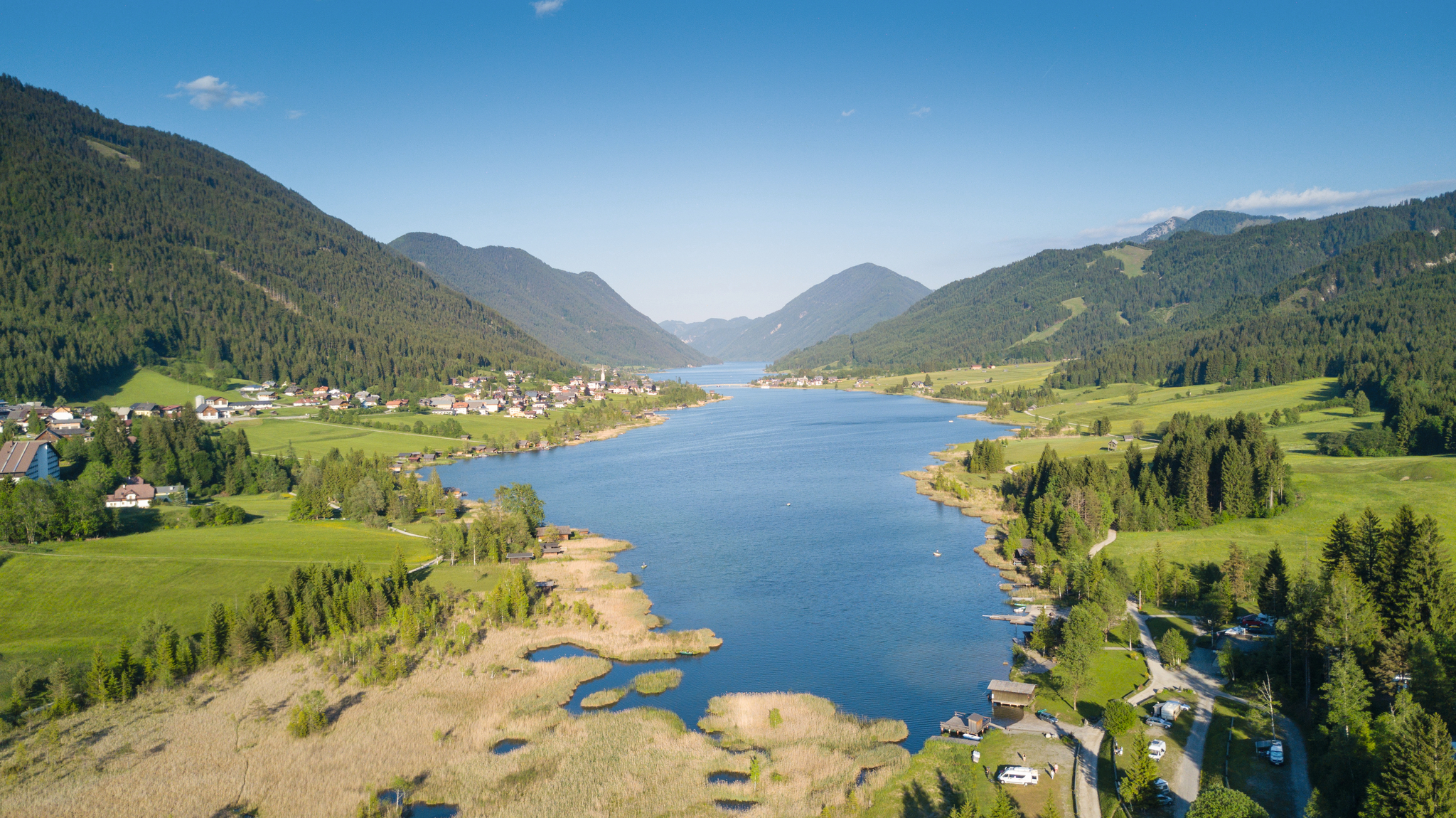 Landschaft mit Bergsee, umgeben von Wiesen und Wäldern unter klarem Himmel.