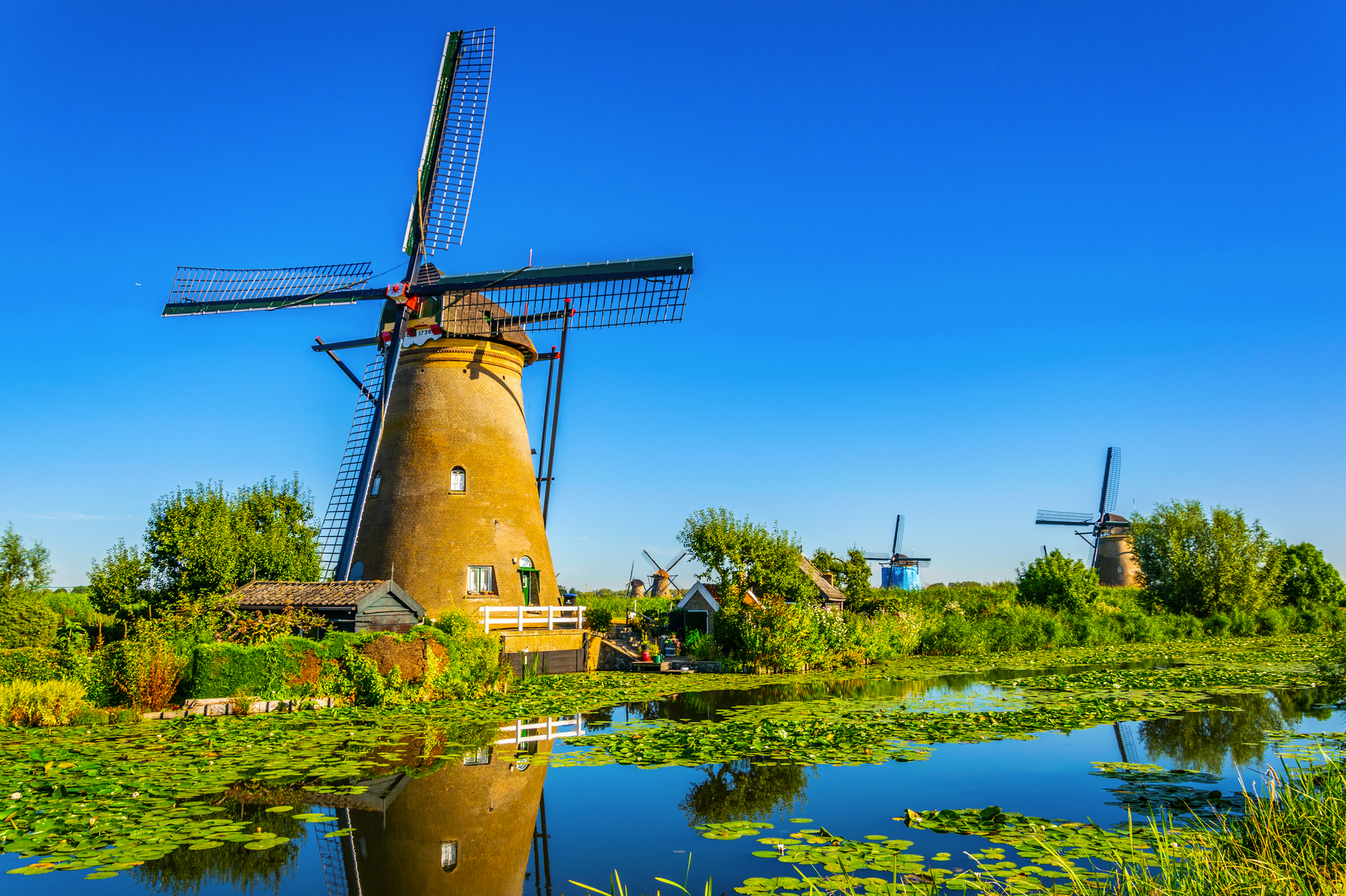 Windmühle am Wasser umgeben von grüner Landschaft und blauem Himmel.