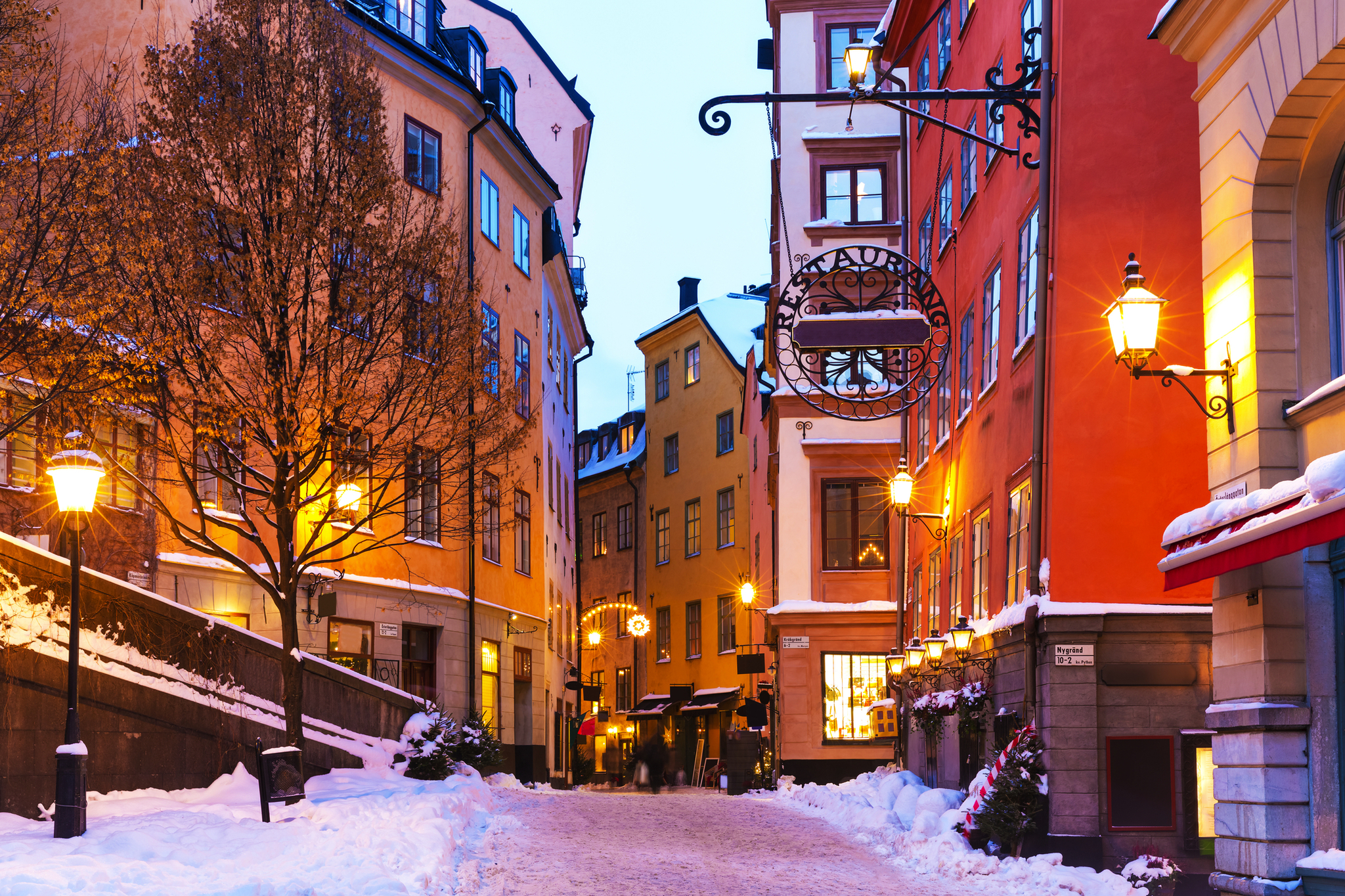 Verschneite Altstadtgasse mit bunten Fassaden und Straßenlaternen bei Abenddämmerung.