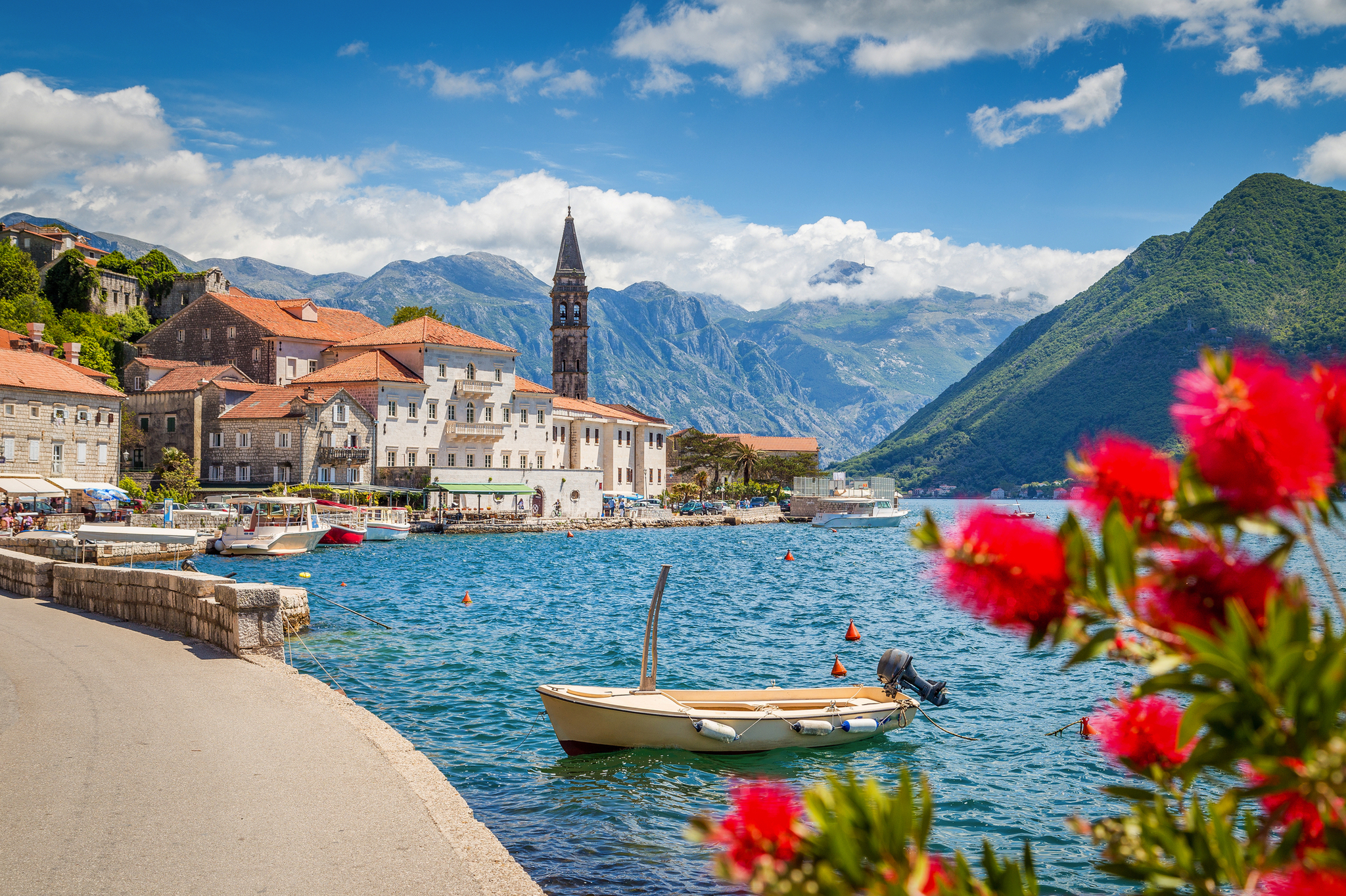 Küstenstadt mit Glockenturm am blauen Meer, umgeben von Bergen und blühenden Blumen.
