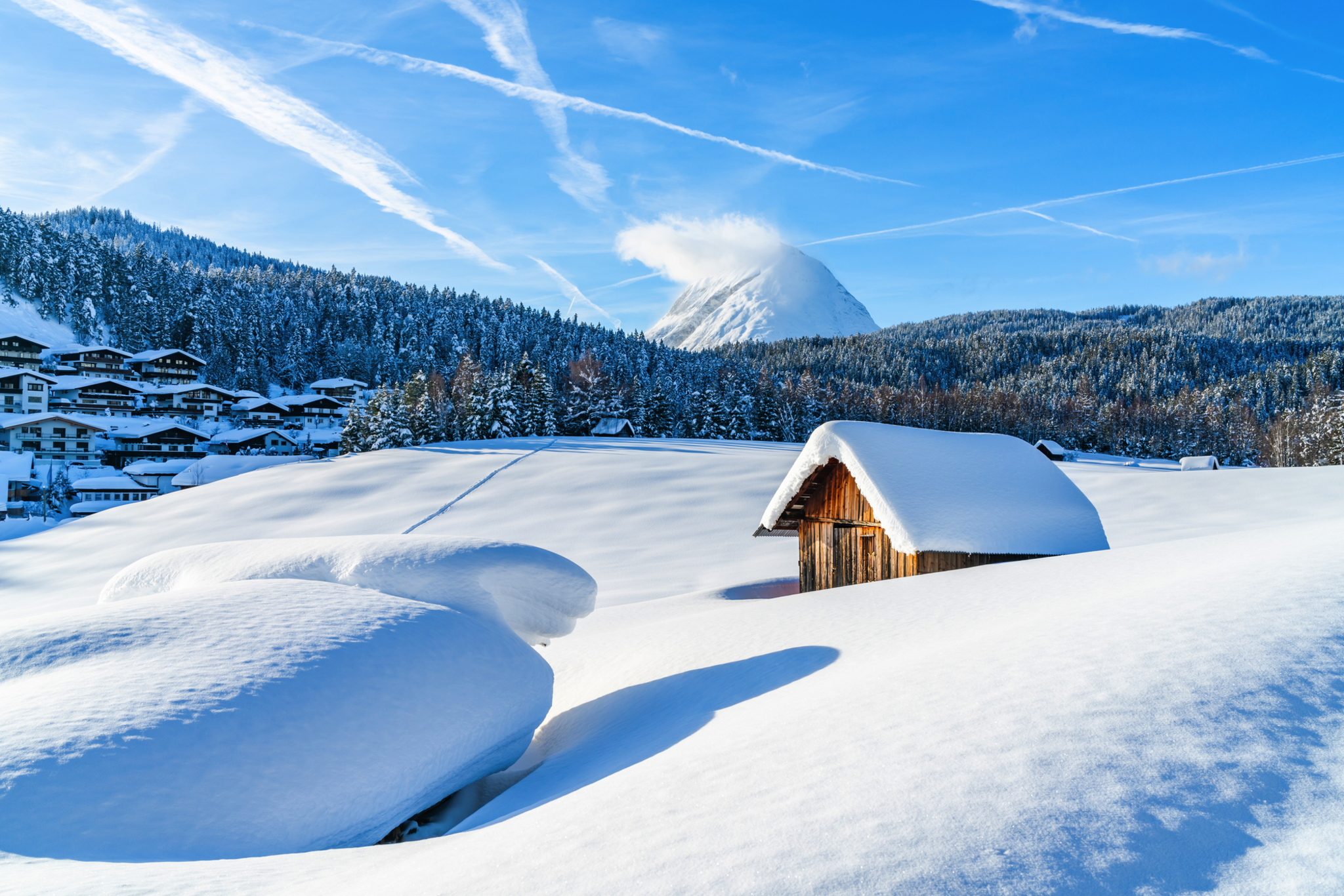 Verschneite Landschaft mit Hütte und Bergen im Hintergrund bei blauem Himmel.