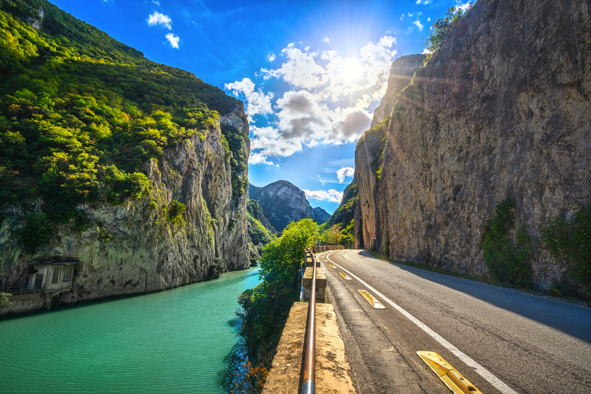 Straße entlang türkisfarbenem Fluss in enger Schlucht unter blauem Himmel mit Sonne.