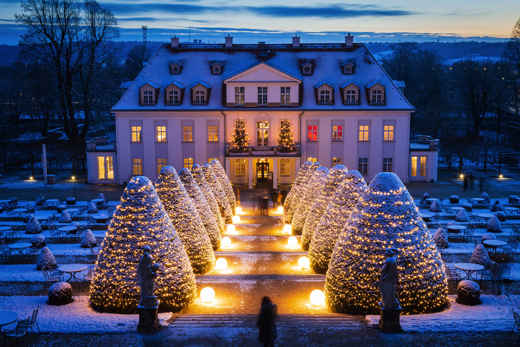 Beleuchtetes Herrenhaus im Winter mit schneebedecktem Garten bei Dämmerung.