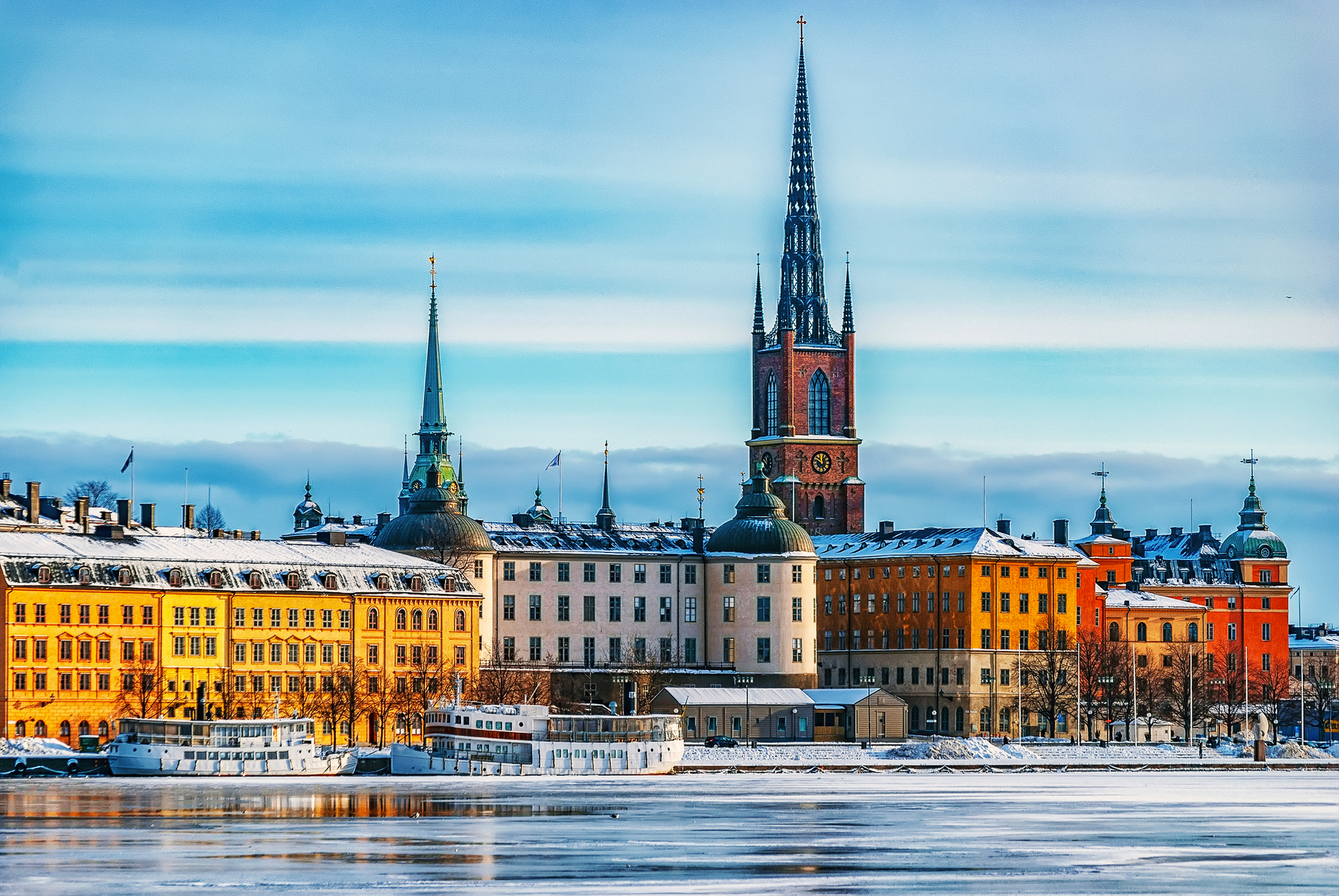 Verschneite Stadtansicht mit bunten Häusern und Kirchtürmen am Wasser.