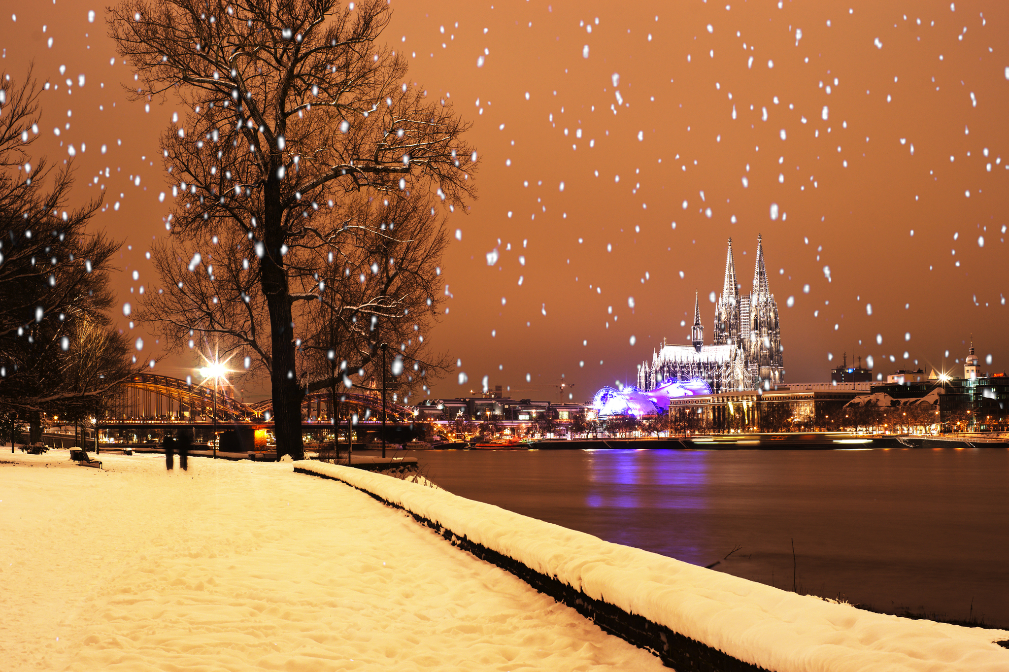 Winterliche Stadtansicht mit schneebedecktem Flussufer und beleuchtetem Dom im Hintergrund.