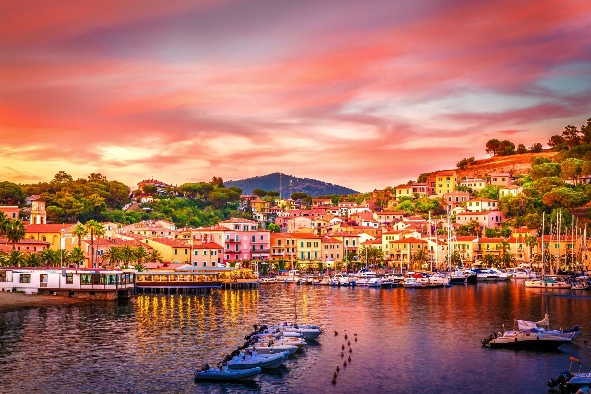 Hafen von Porto Azzurro auf der Insel Elba bei Sonnenuntergang mit Booten im Wasser und bunten Gebäuden im Hintergrund.