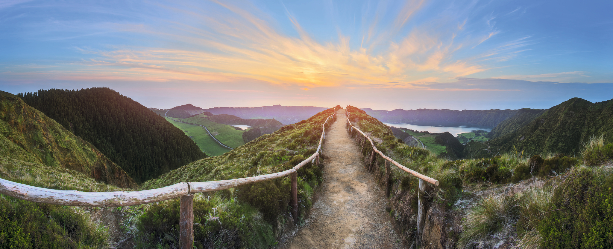 Pfad mit Holzgeländer führt durch grüne hügelige Landschaft bei Sonnenuntergang.