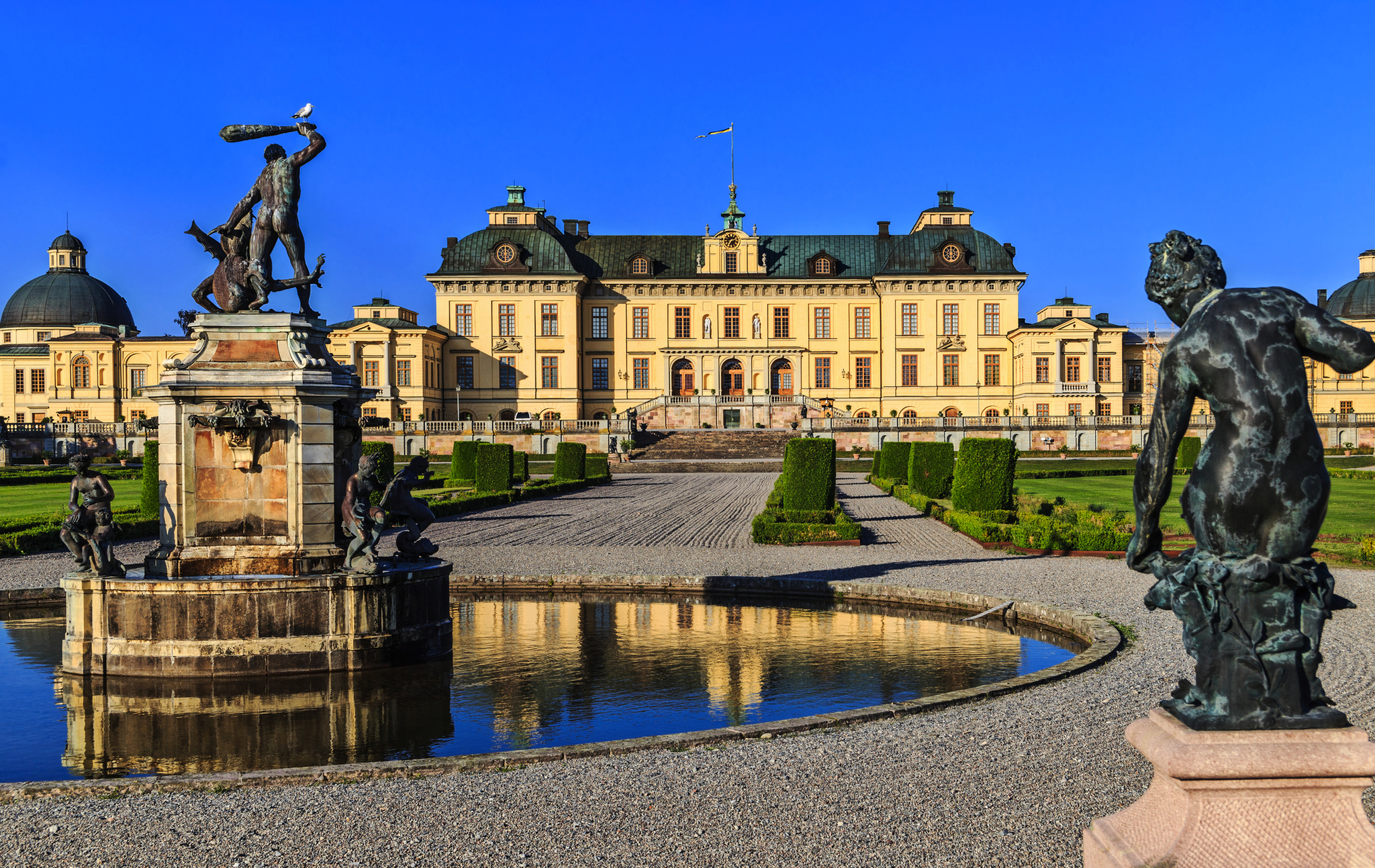 Schloss Drottningholm mit Gartenanlage und Statuen im Vordergrund an einem sonnigen Tag.