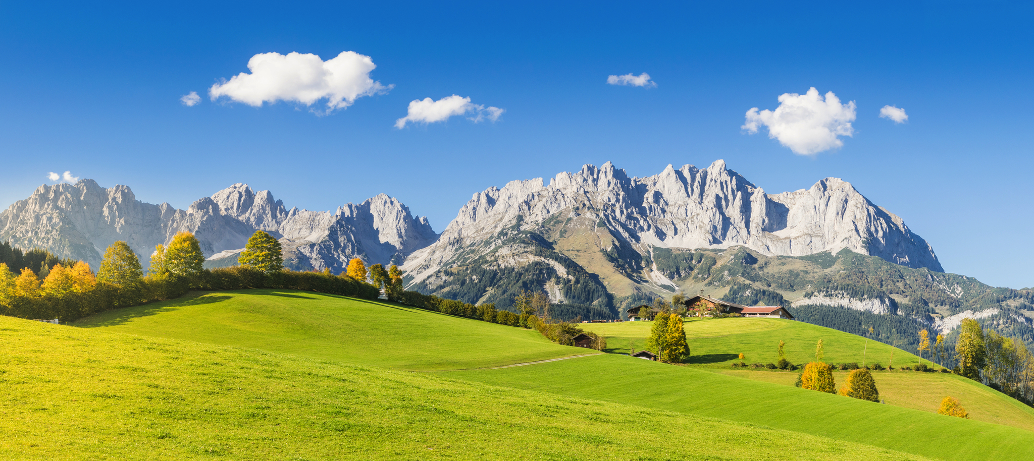 Grüne Wiesen vor einer Berglandschaft mit klarem Himmel und vereinzelten Wolken.