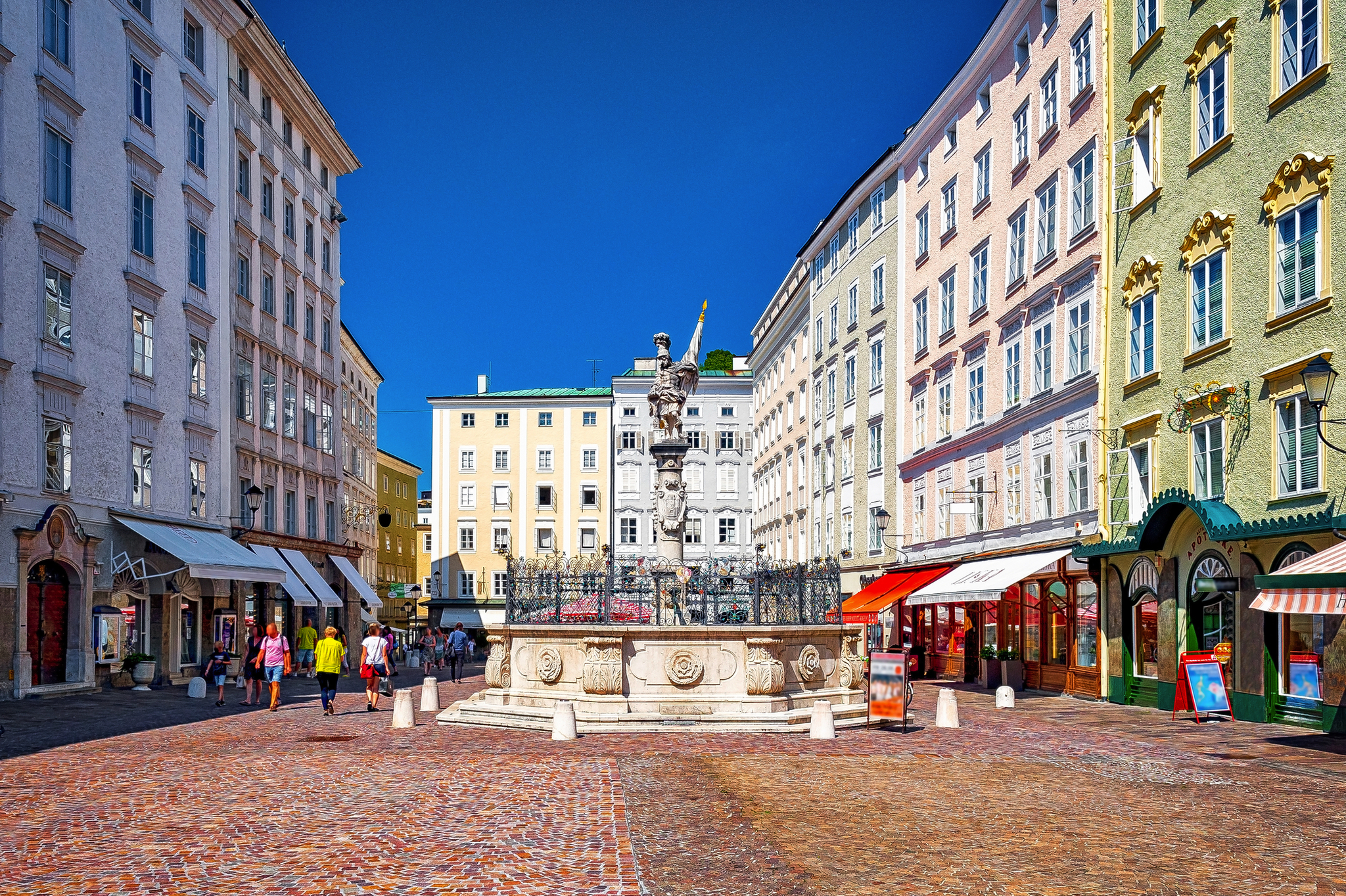 Städtischer Platz mit Springbrunnen und umliegenden Pastellgebäuden bei klarem Himmel.