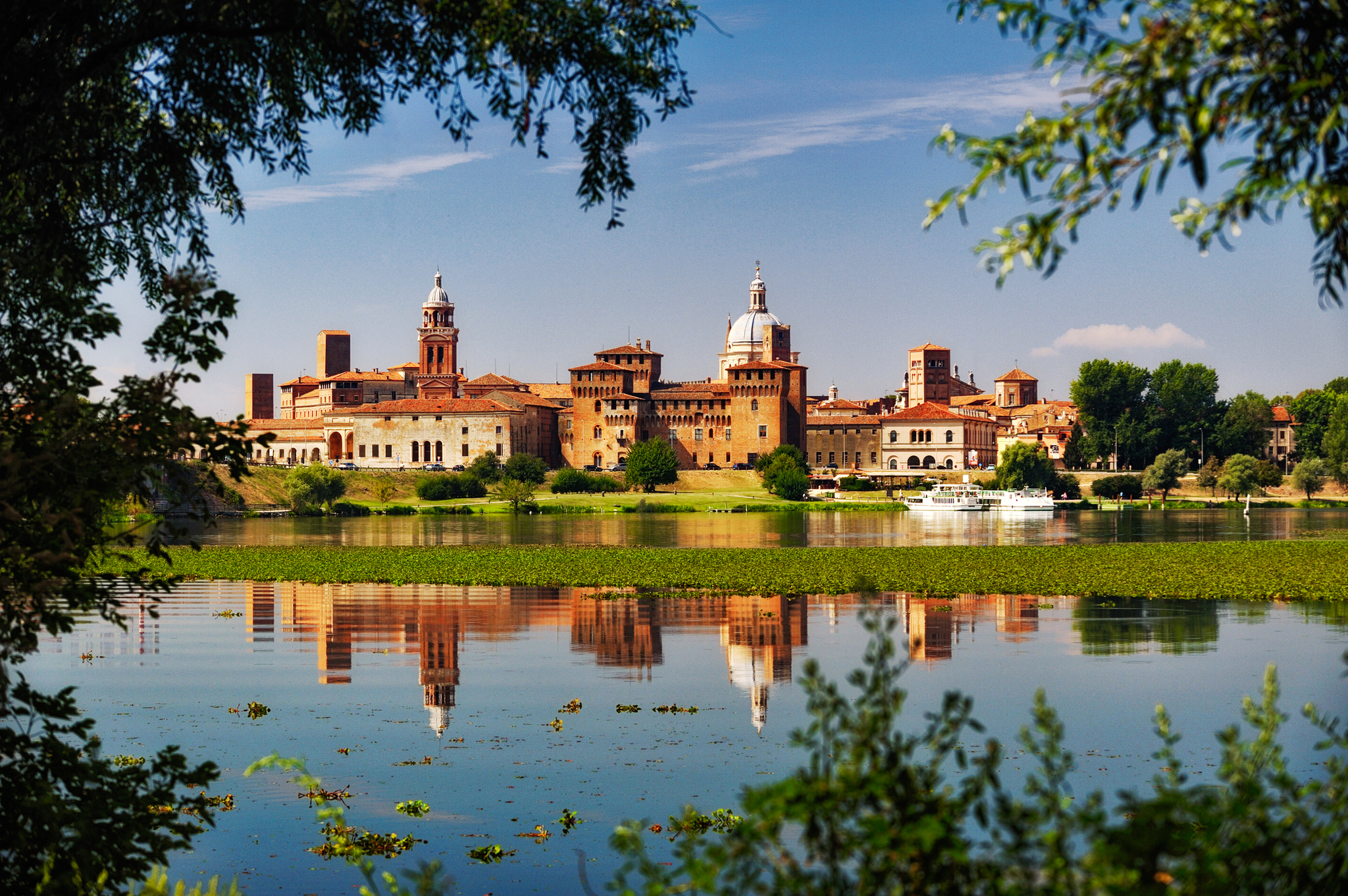 Historische Stadt mit Wasserreflektion und Bäumen im Vordergrund.