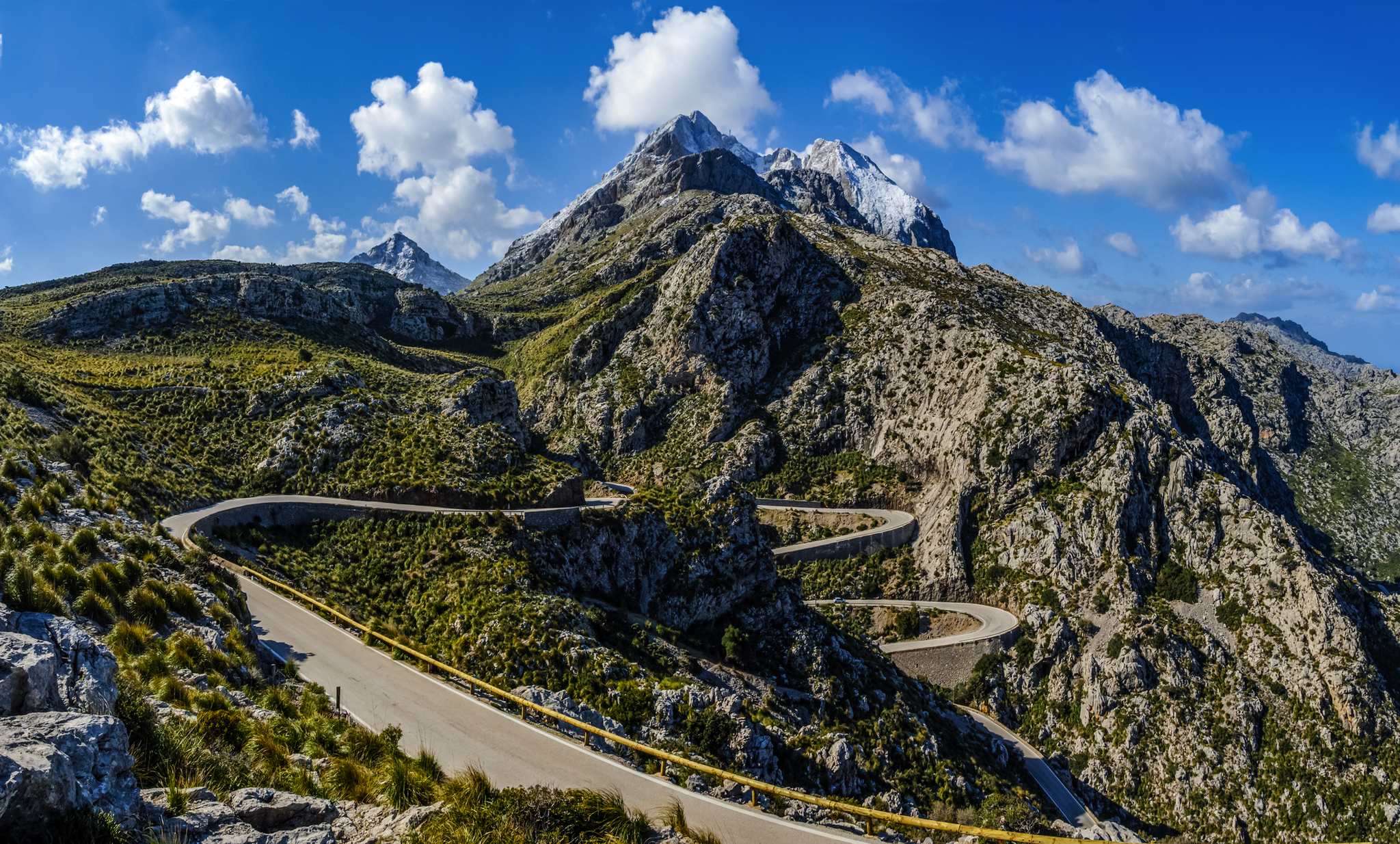 Serpentinenstraße in bergiger Landschaft mit blauem Himmel und Wolken.