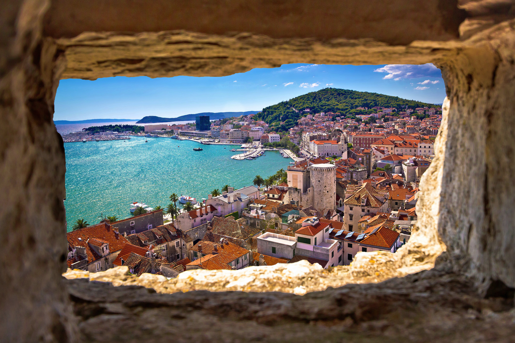 Blick durch Steinfenster auf Küstenstadt mit Meer und Hügel im Hintergrund.