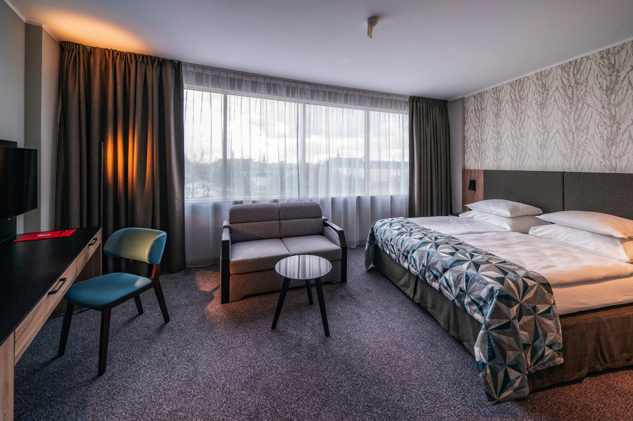 Hotelzimmer mit Doppelbett, Sofa, Tisch, Stuhl und Schreibtisch vor einem großen Fenster.
