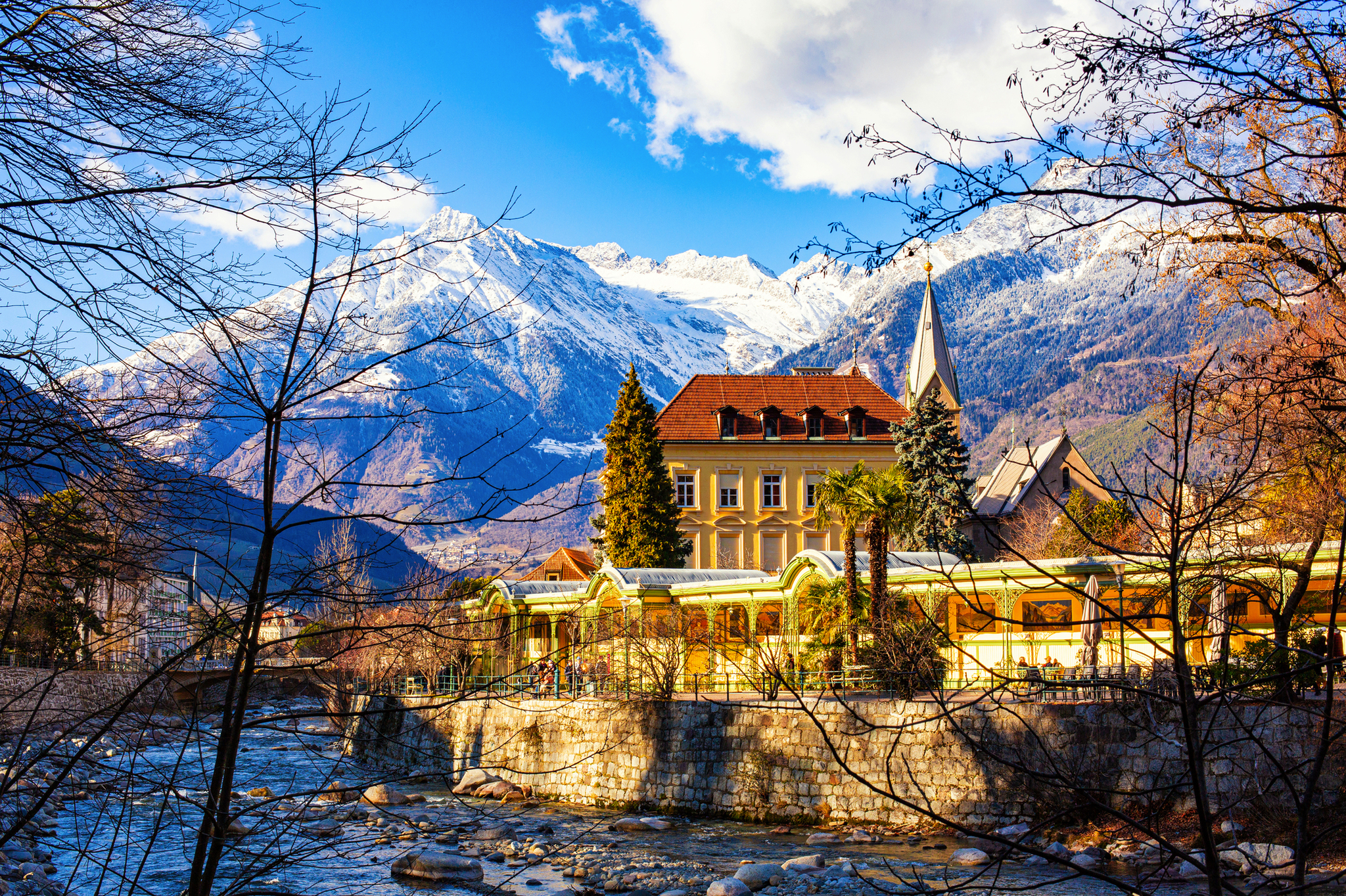 Großes Gebäude mit Turm vor schneebedeckten Bergen, umgeben von Bäumen und Fluss.