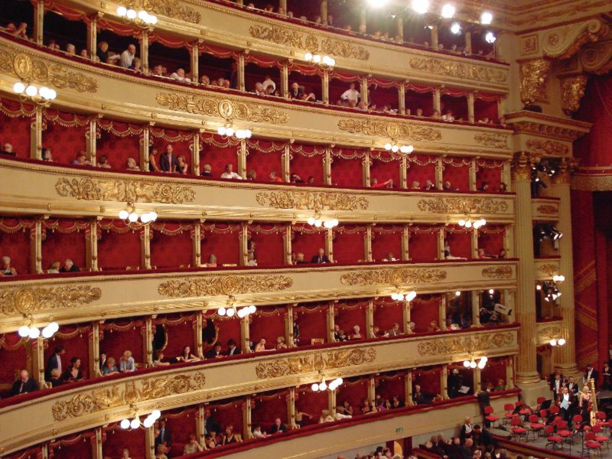 Innenansicht eines Theaters mit roten Logen und Zuschauern.