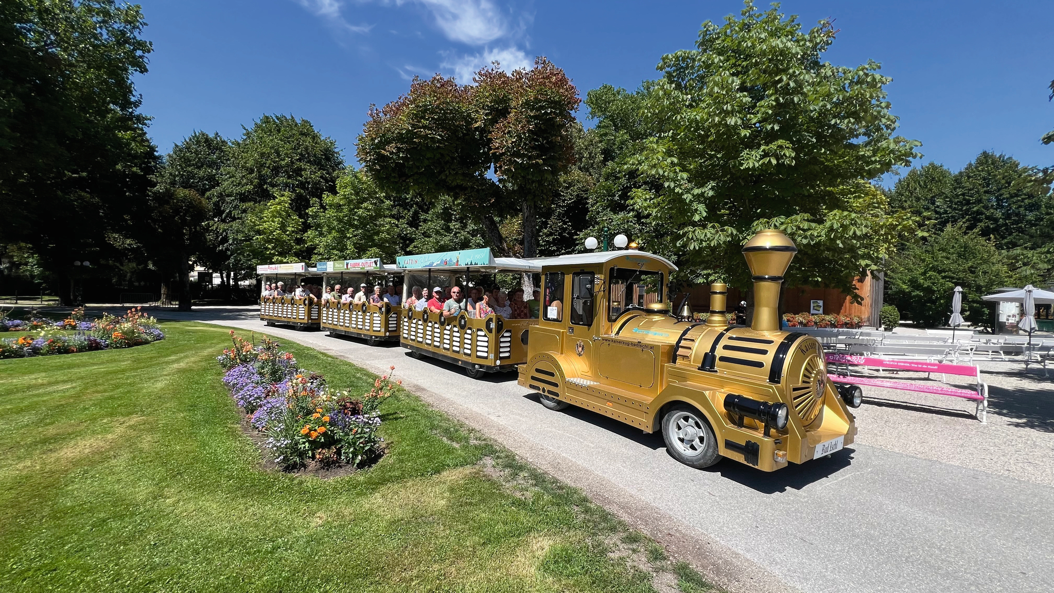 Kleiner goldener Touristen-Zug fährt durch einen Park bei sonnigem Wetter.