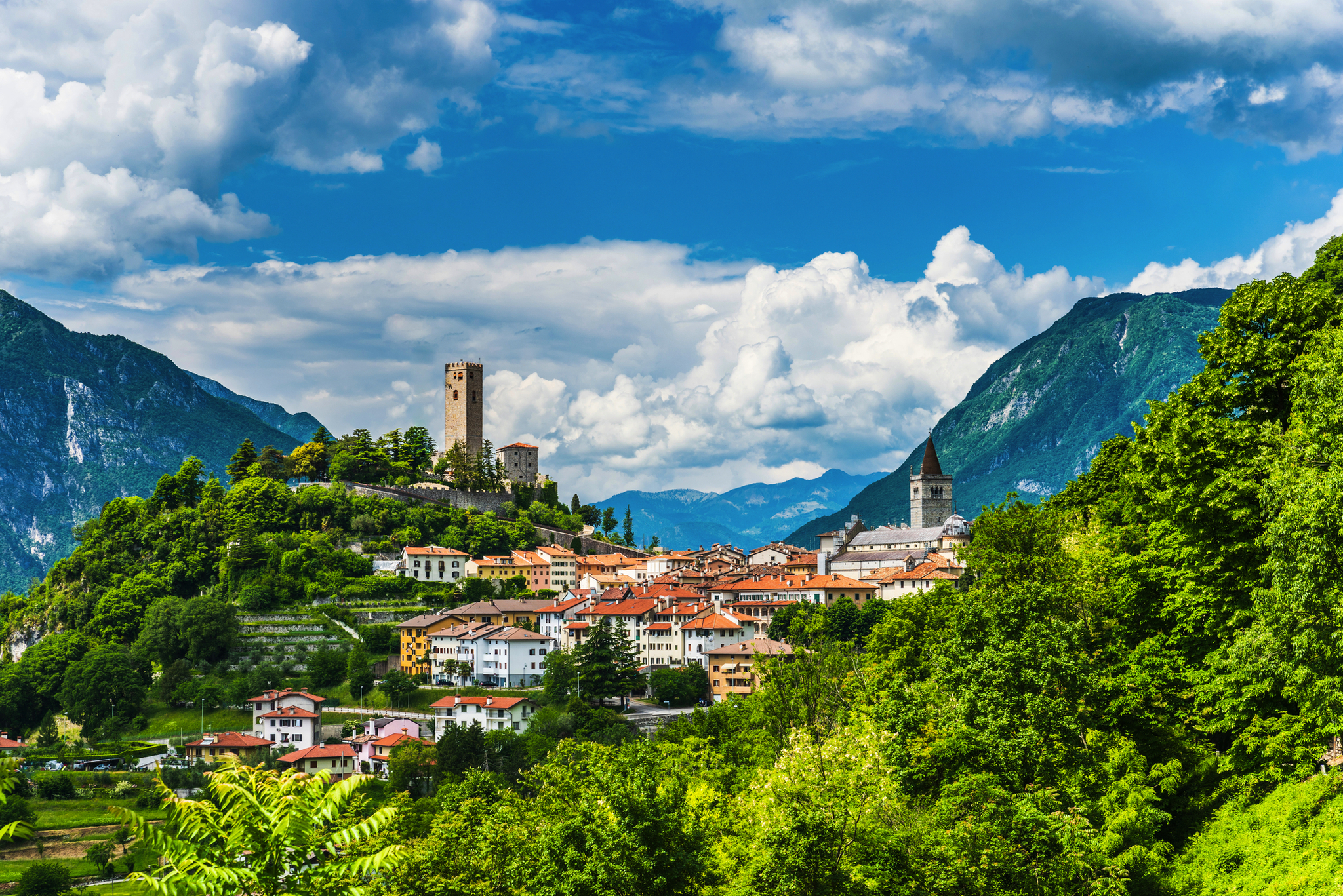 Hügelige Landschaft mit Dorf und alter Festung vor bewaldeten Bergen und blauem Himmel.