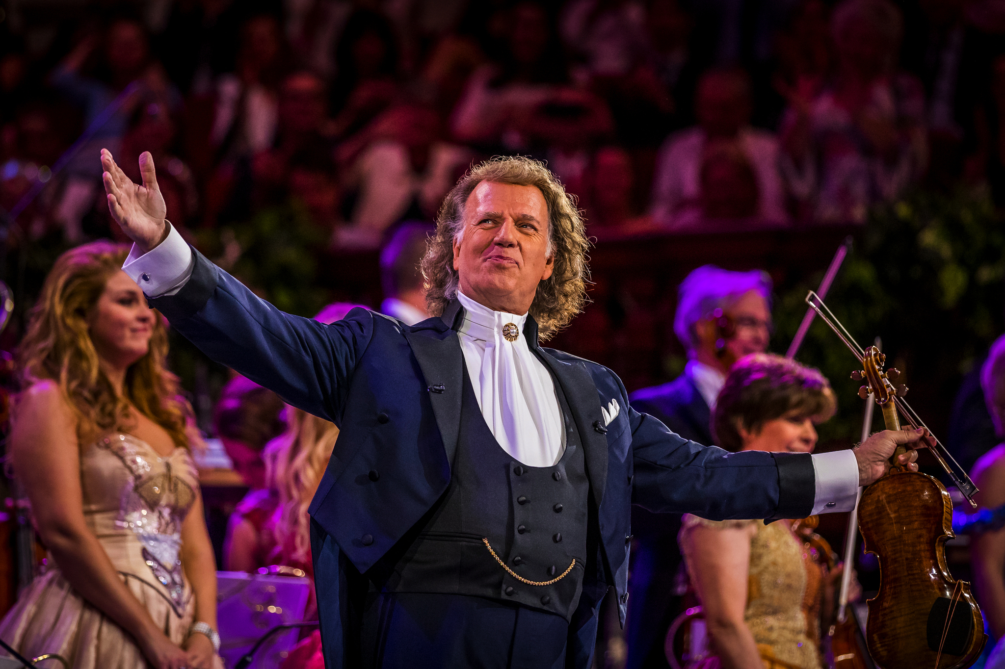 Ein Dirigent in festlicher Kleidung dirigiert ein Orchester auf einer Bühne.