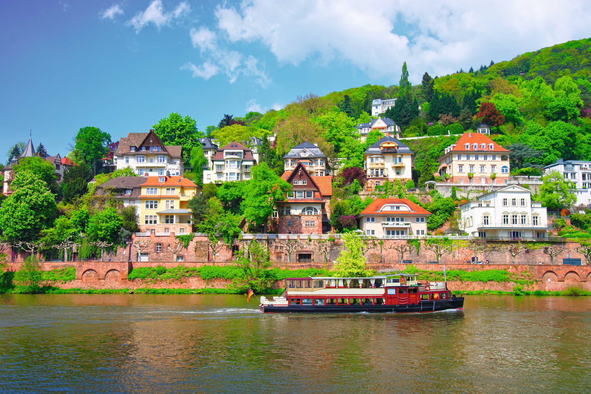 Schiff auf dem Neckar in Heidelberg, im Hintergrund historische Gebäude und bewaldete Hügel.