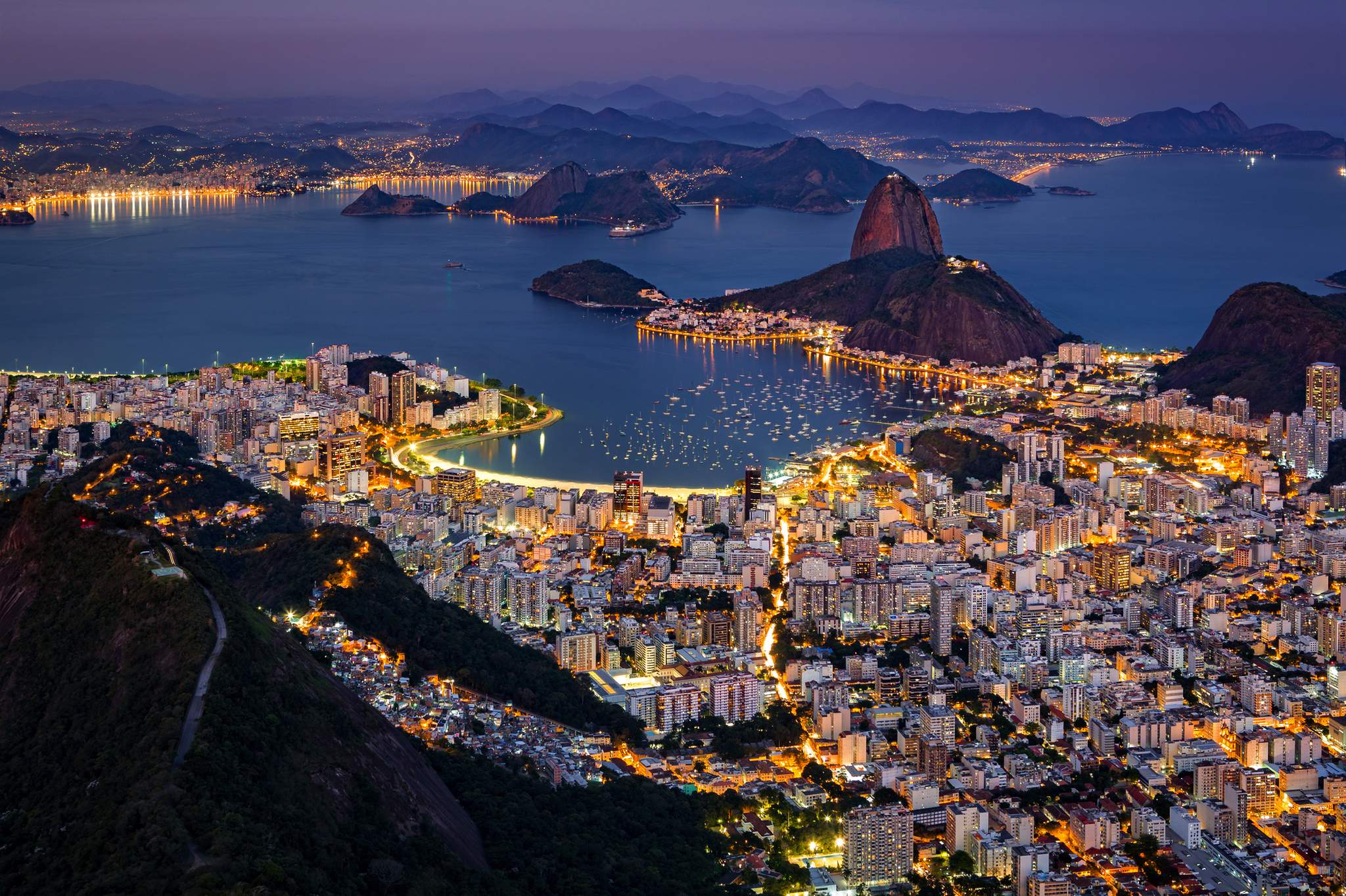 Abendliche Stadtansicht mit beleuchteter Skyline und Küste, Hügel im Hintergrund.